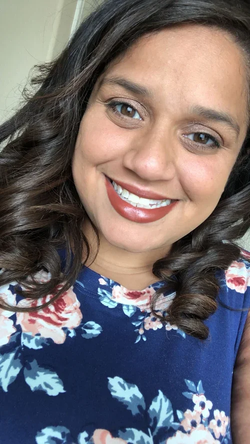 Close-up of a smiling woman with dark, curly hair wearing a blue floral top.