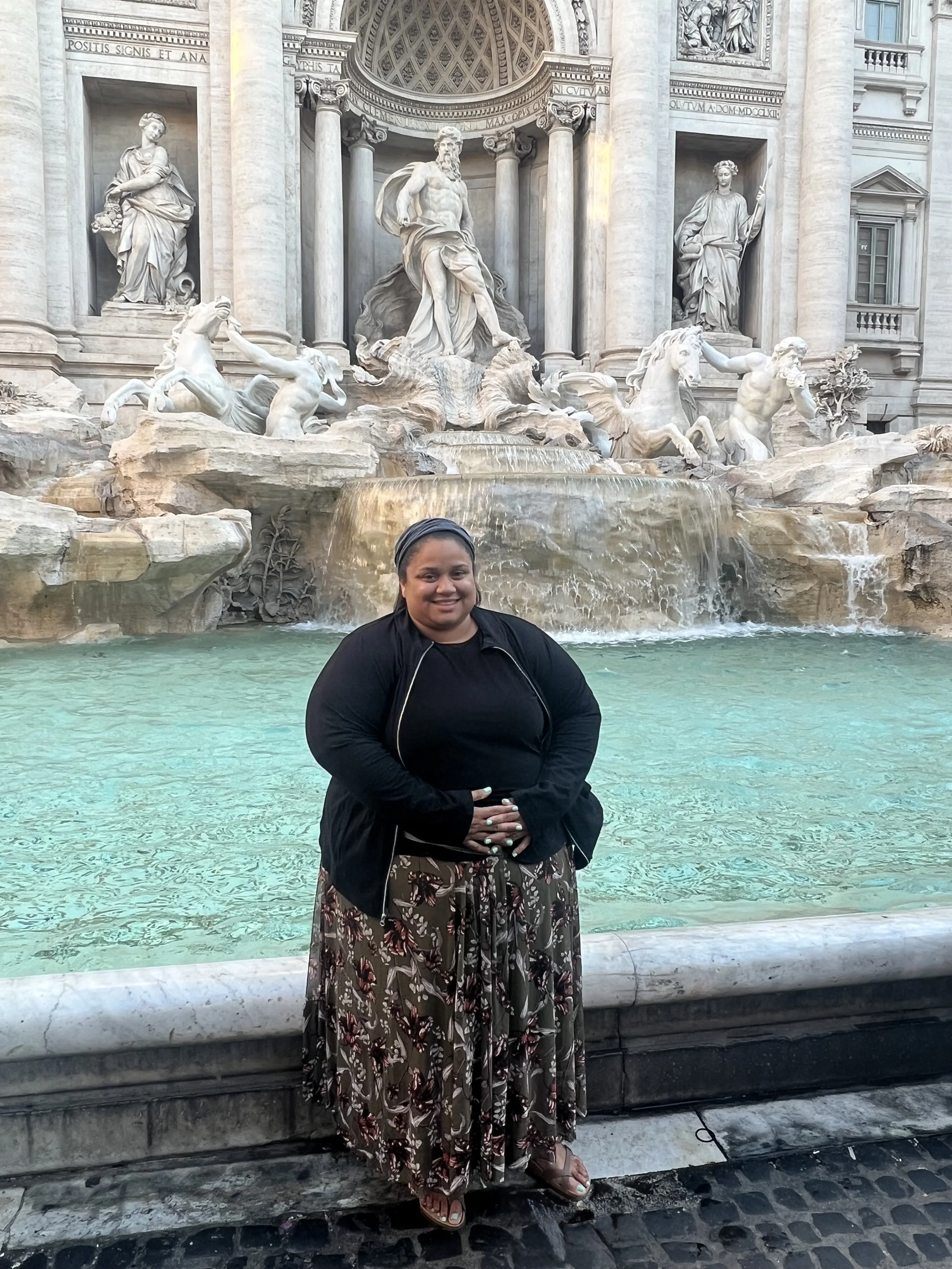 A woman standing in front of the trevi fountain in Rome, Italy, smiling at the camera, wearing a black jacket, patterned skirt, and sandals.