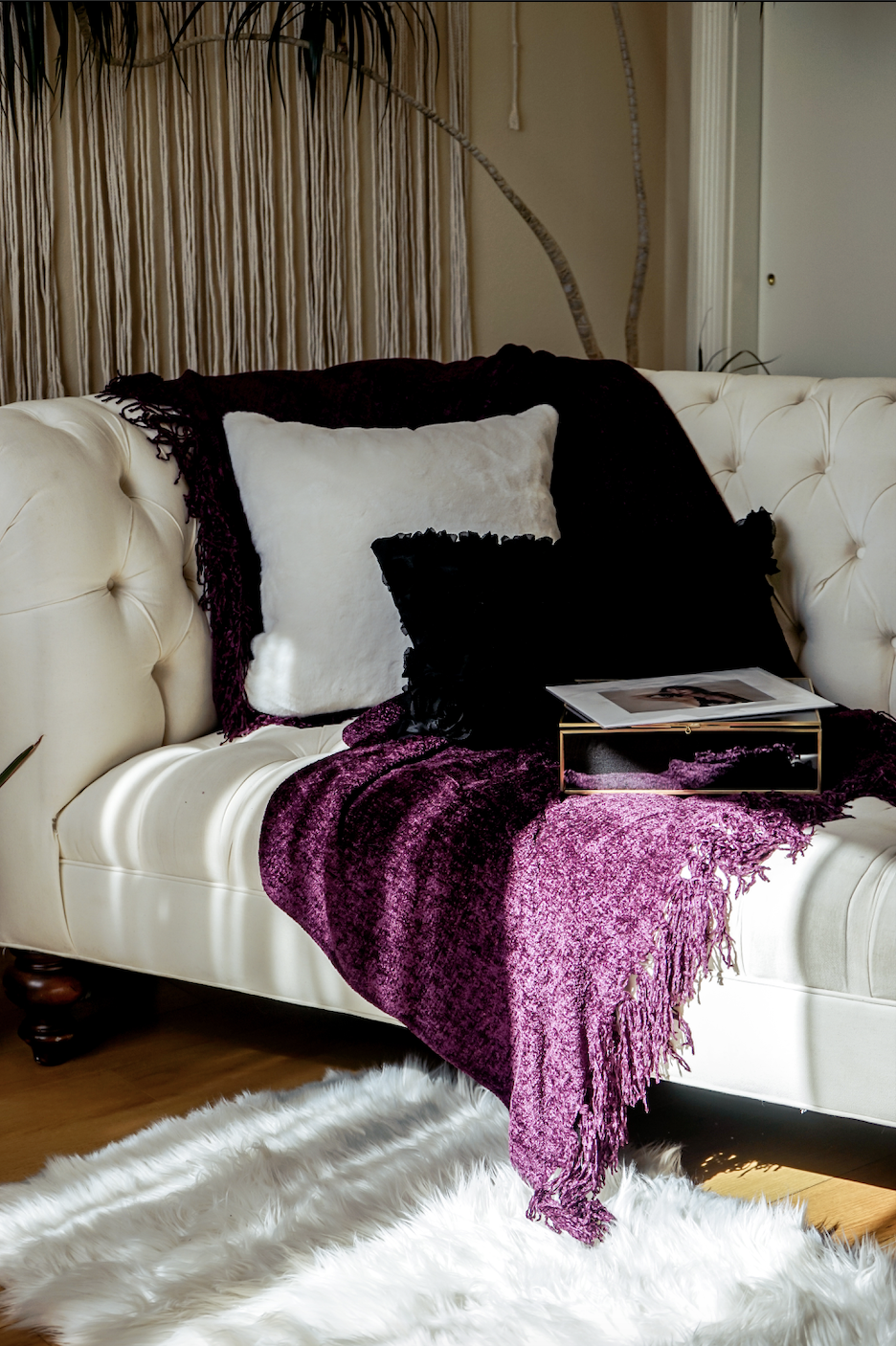 White tufted sofa with purple throw blanket and black and white pillows, next to a magazine on a fluffy rug in a cozy room.