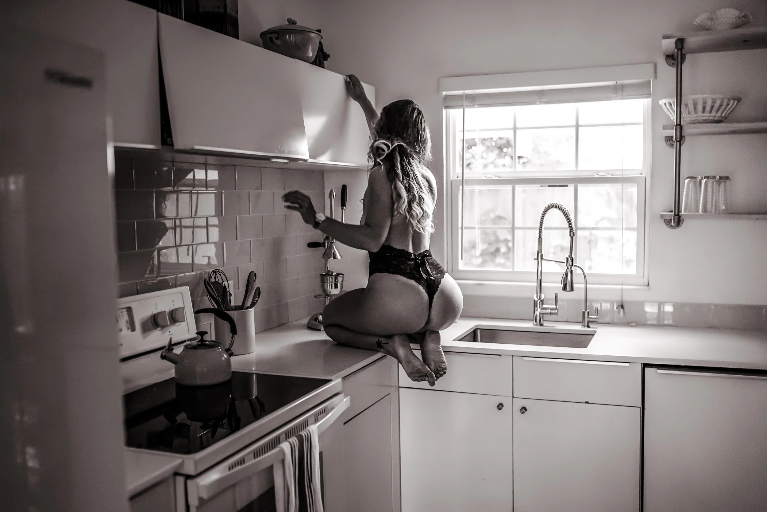 Woman in lingerie on kitchen countertop, reaching for cabinet. Kitchen boudoir photoshoot ideas. Housewife themed photoshoot session. 