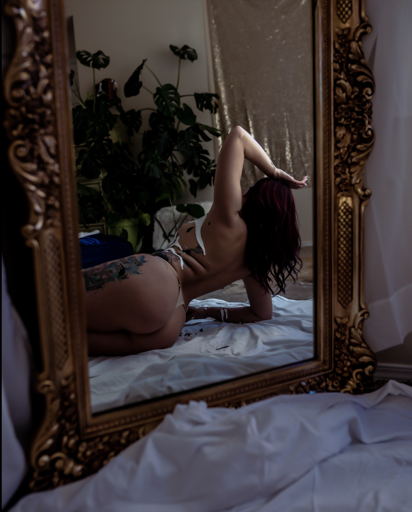 Reflection of woman lying on white sheet in ornate mirror with plants in background. Creative and messy intimate portrait session to connect with our wild feminine and authentic self