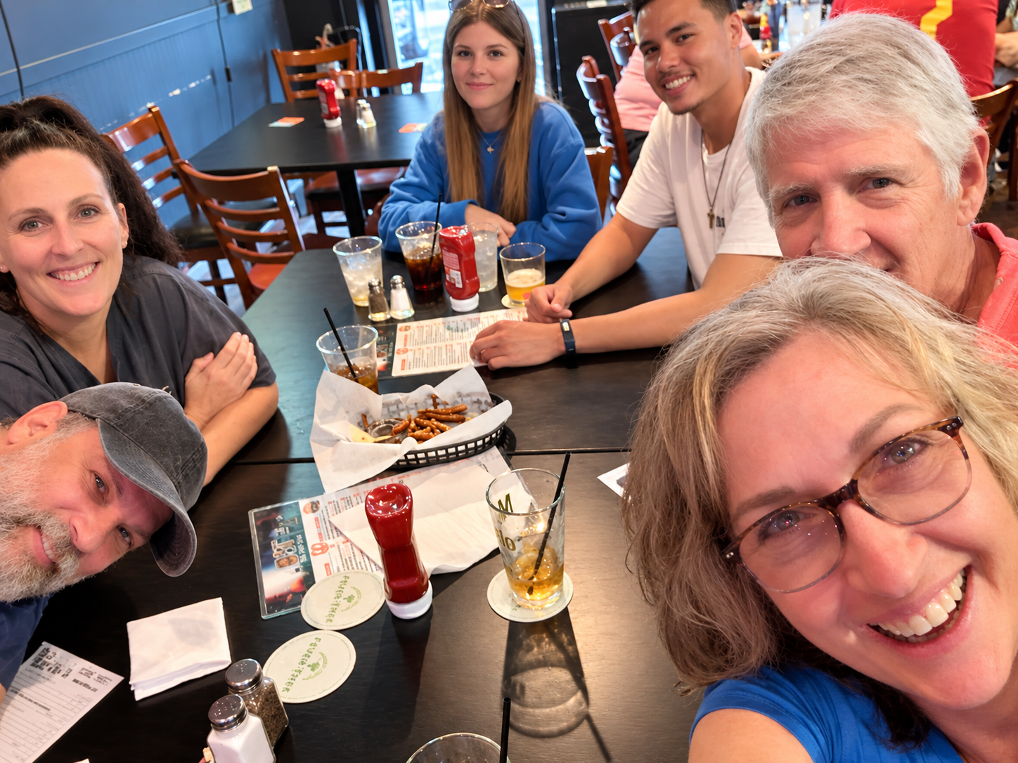 Group of seven people smiling and gathering around a table in a restaurant, with food and drinks, including pretzels, soda, and beer, on the table.