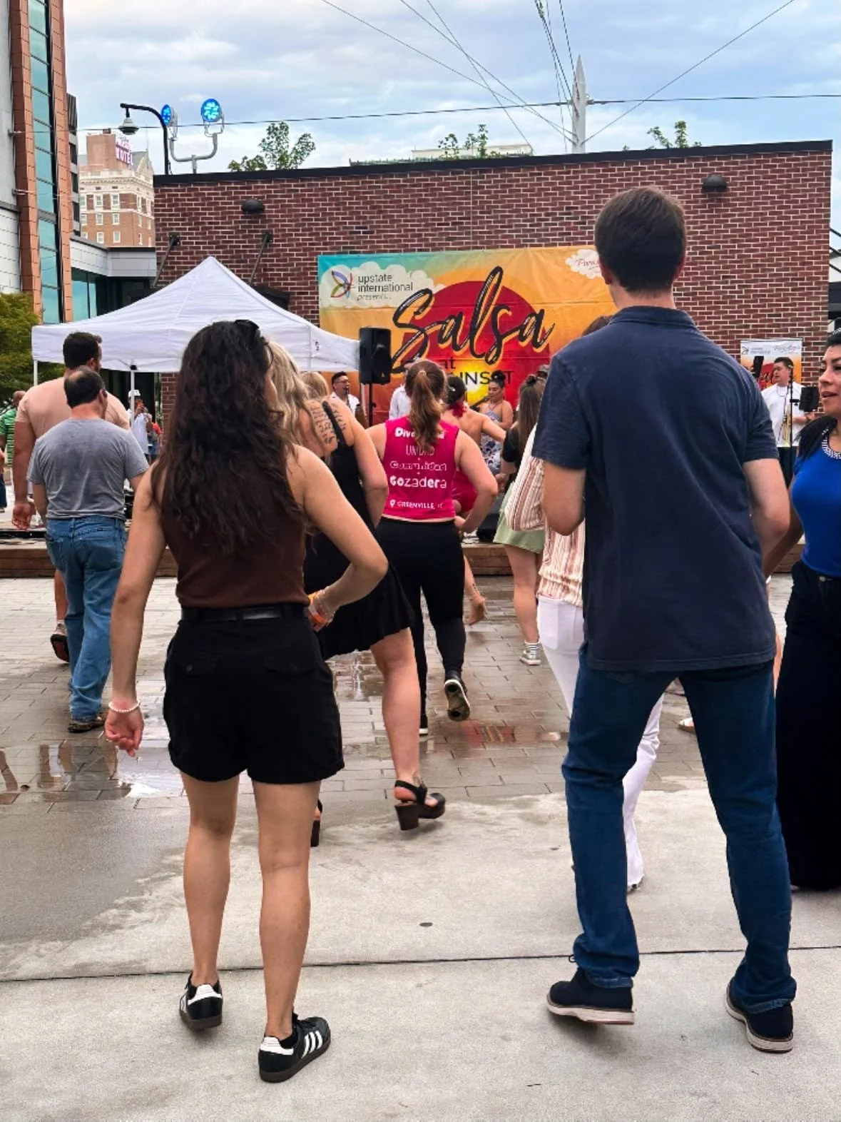 People gather outdoors in front of a stage with a colorful banner that says 'Salsa at Main St.' and a tent, with a brick building in the background.