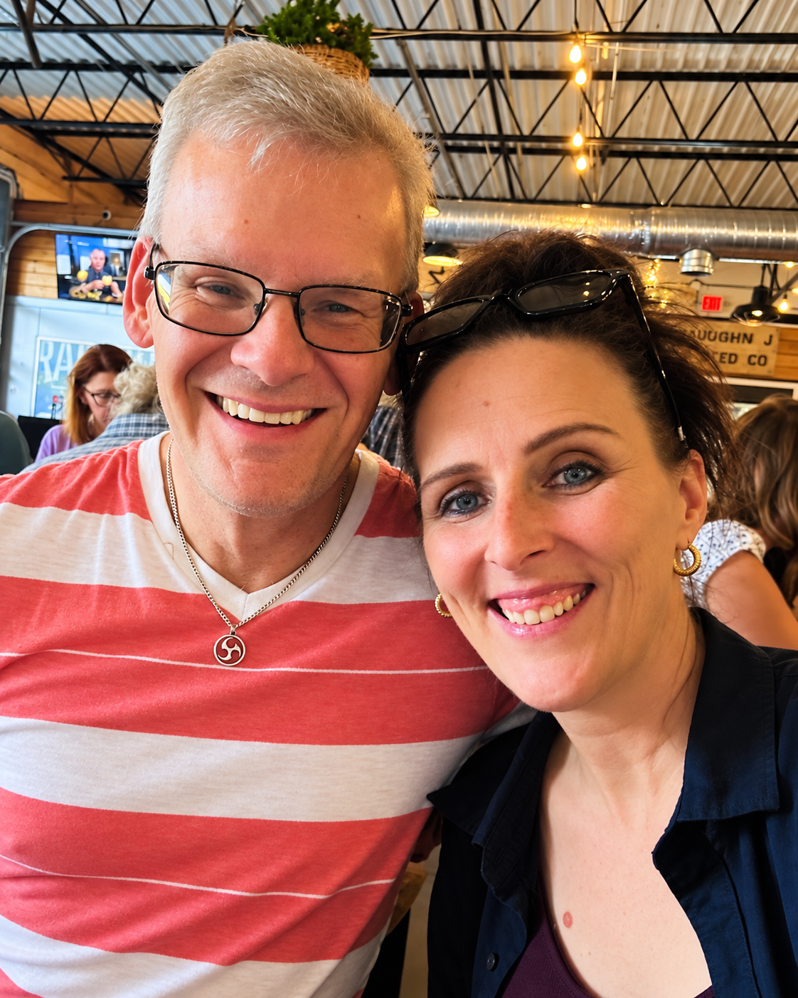 A smiling man with glasses and a woman with glasses on her head in a restaurant or cafe setting.