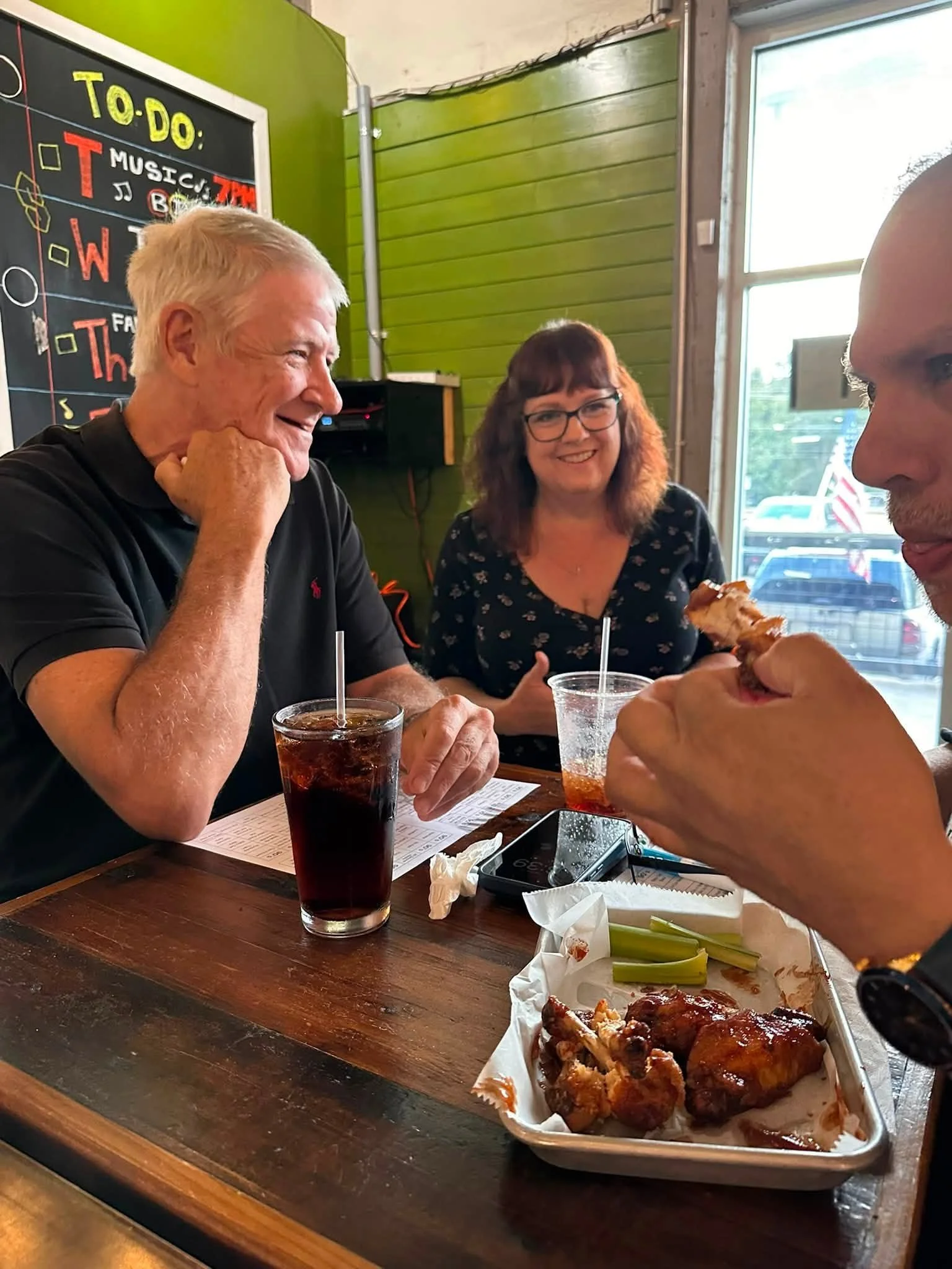 Three people sitting at a wooden table in a restaurant or cafe, enjoying fried chicken and drinks. The background includes a chalkboard menu and large windows letting in natural light.