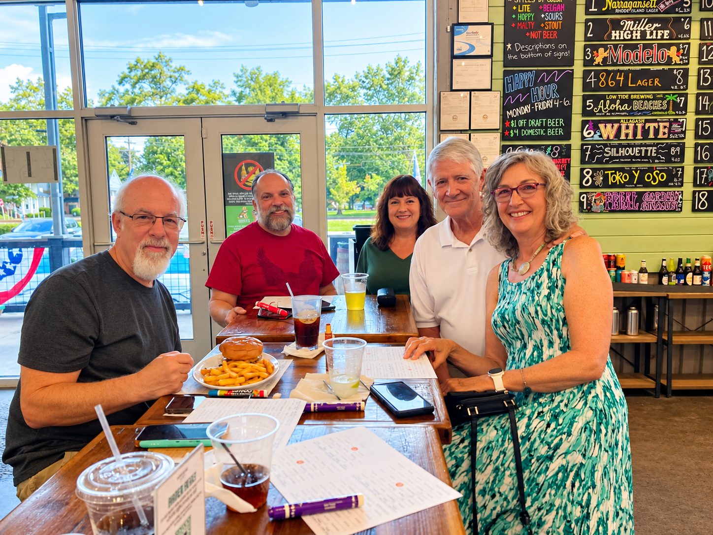 Five adults smiling and enjoying food and drinks at a table inside a casual restaurant with a colorful menu board on the wall. Outside the restaurant window, trees and a parking lot are visible.