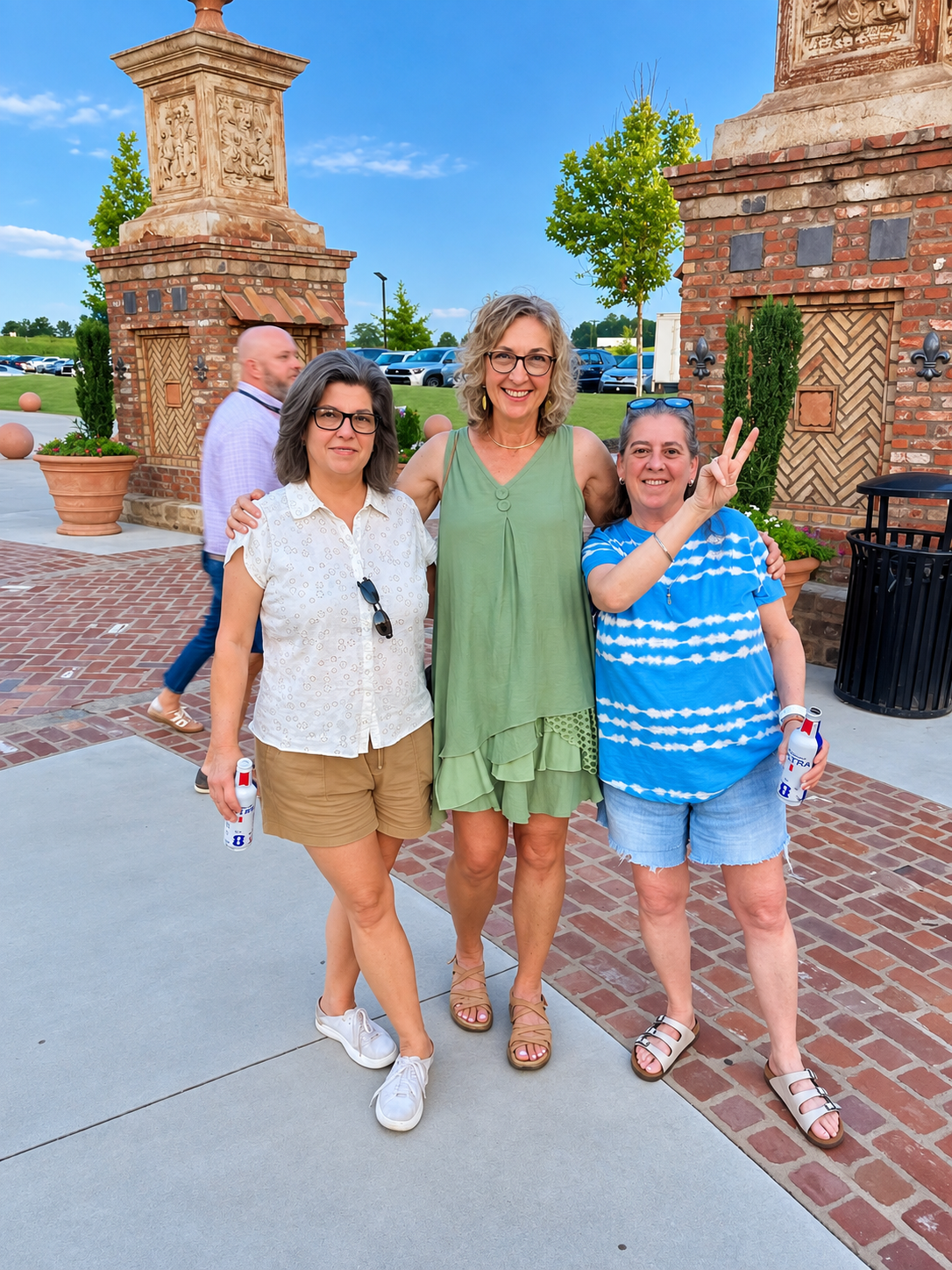Three women standing outdoors by brick structures during a sunny day, with two holding drinks and one making a peace sign, and a man walking behind them.