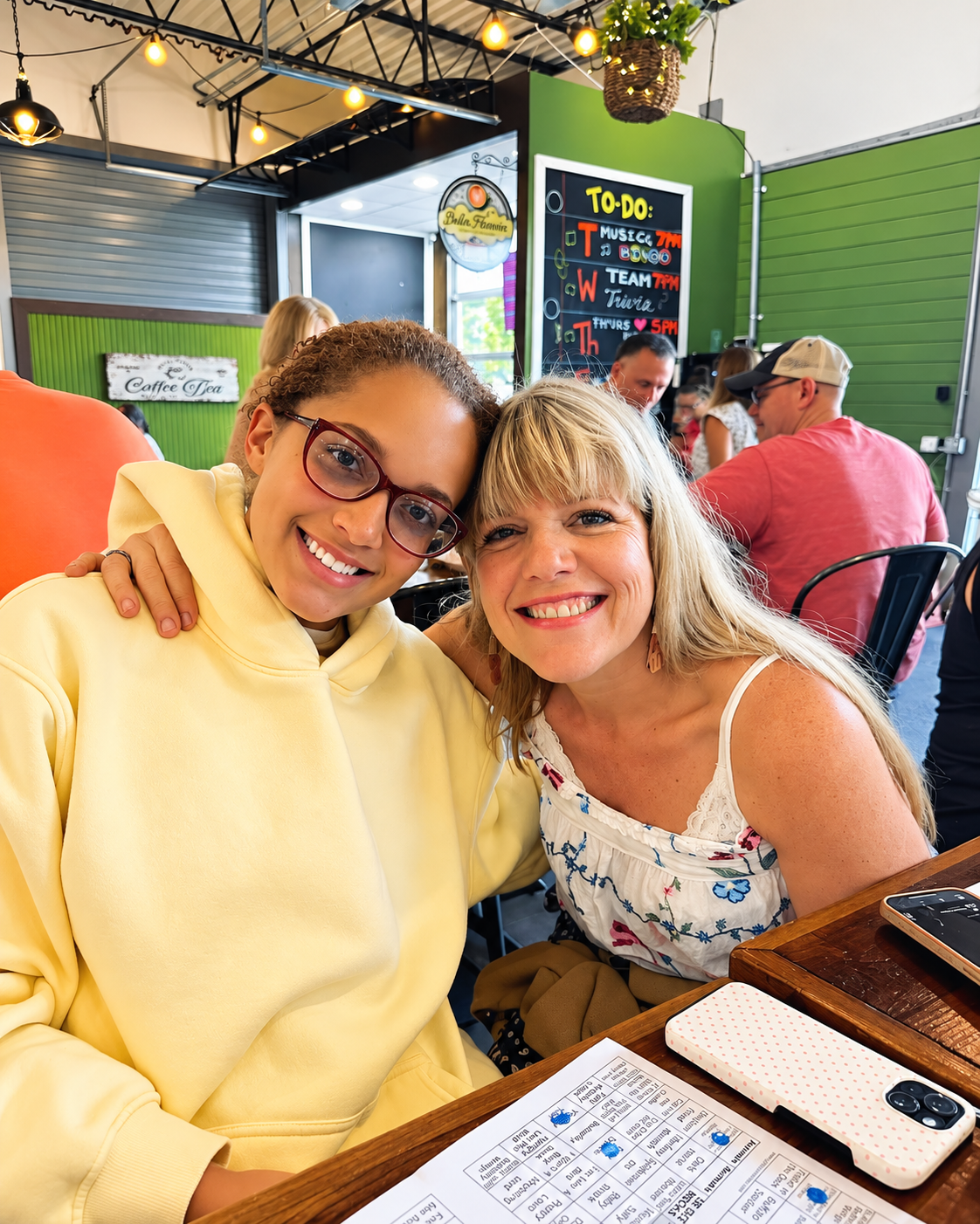 Two women smiling and sitting closely together at a restaurant table, with the younger woman wearing glasses and a yellow sweatshirt and the older woman in a white floral top. There are other people in the background and a chalkboard menu with a to-d