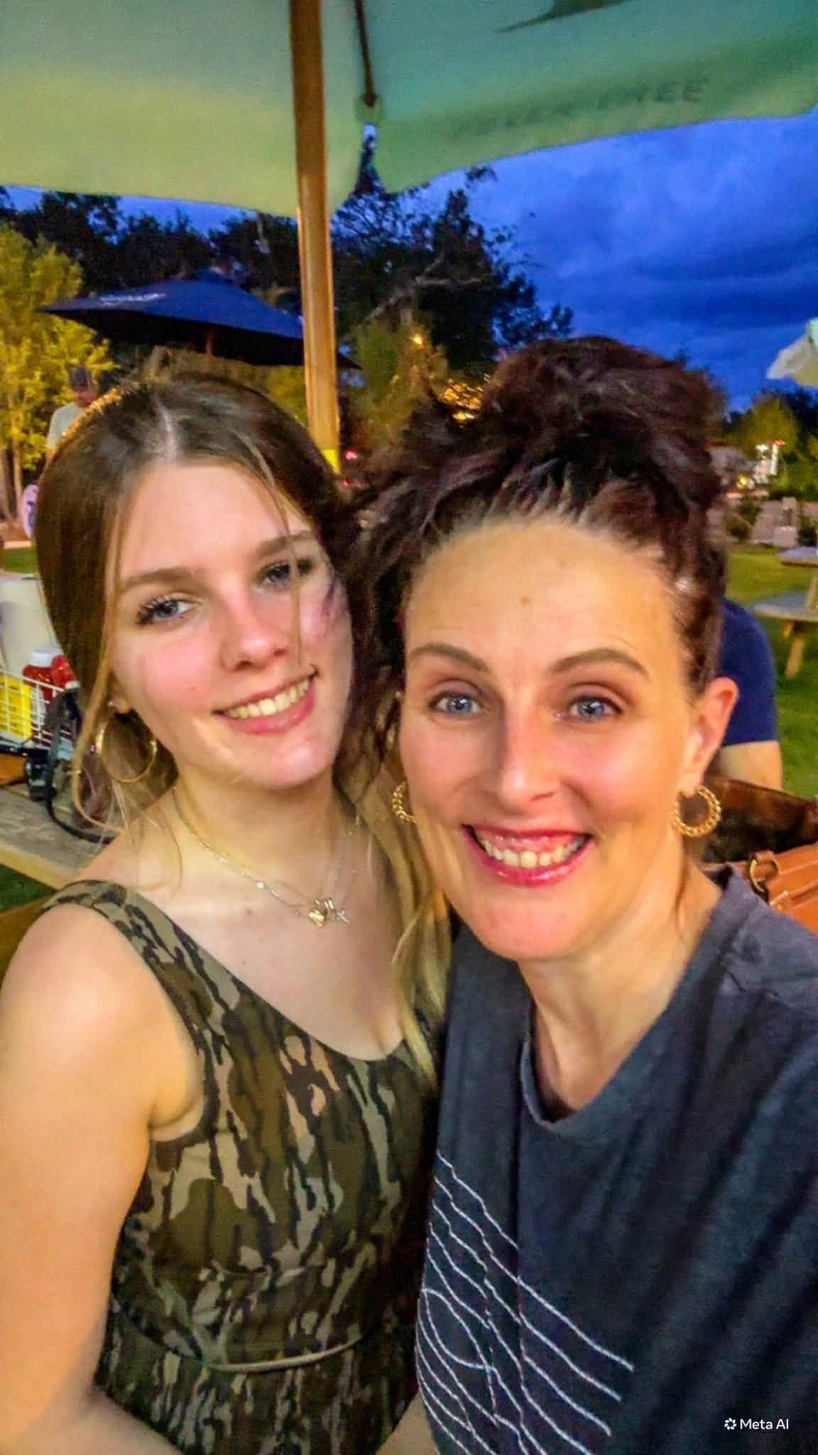 Two women smiling for a selfie outdoors at night with trees, umbrellas, and people in the background.