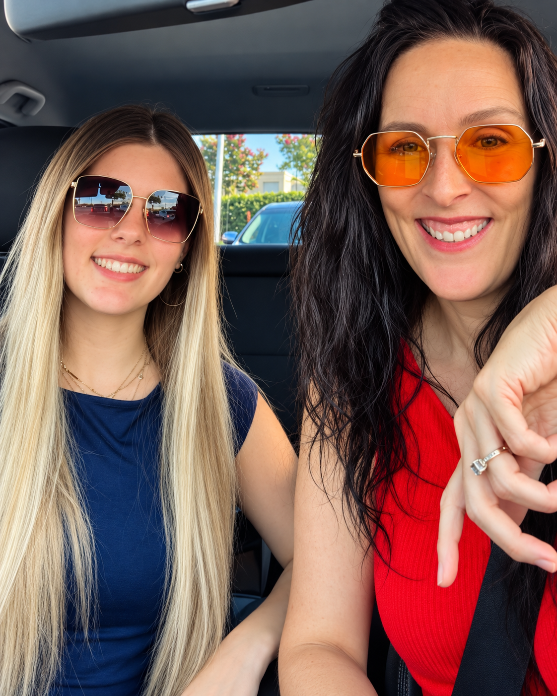 Two women sitting in a car, smiling for a selfie. They are wearing sunglasses, and have long hair. The woman on the left has blonde hair and is wearing a dark blue top. The woman on the right has curly dark hair, is wearing a red top, and has a ring 