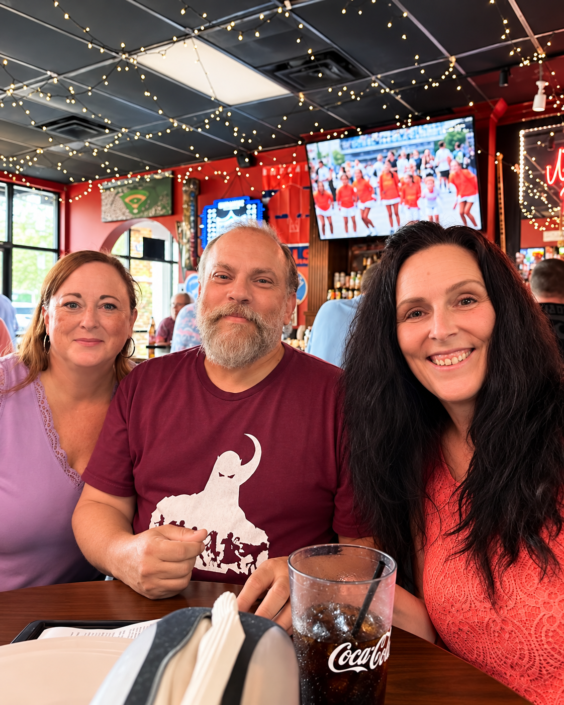 Three people sitting together at a restaurant table, smiling at the camera. The restaurant has string lights, a TV showing a cheerleading team, and a bar in the background. A drink with a straw is on the table.