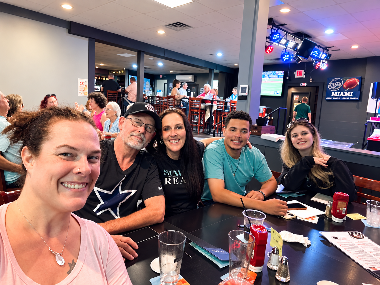 Group of five friends sitting at a restaurant table in a sports bar, smiling for the camera with drinks and condiments on the table, with a sports game playing on TV screens in the background.