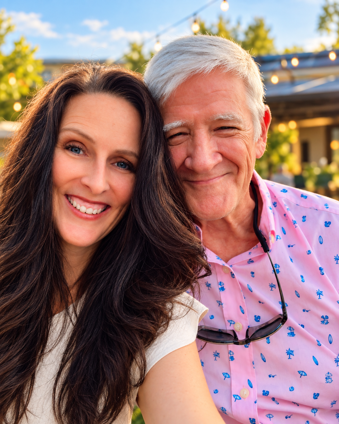 A smiling young woman with long dark hair and blue eyes taking a selfie with an older man with white hair, wearing a pink shirt with blue patterns and sunglasses hanging from his collar, outdoors on a sunny day.