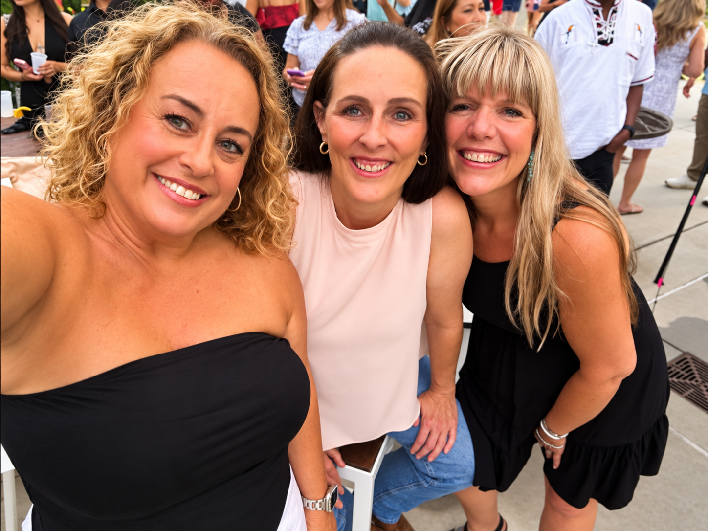 Three women smiling and taking a selfie together at an outdoor event, with other people in the background.