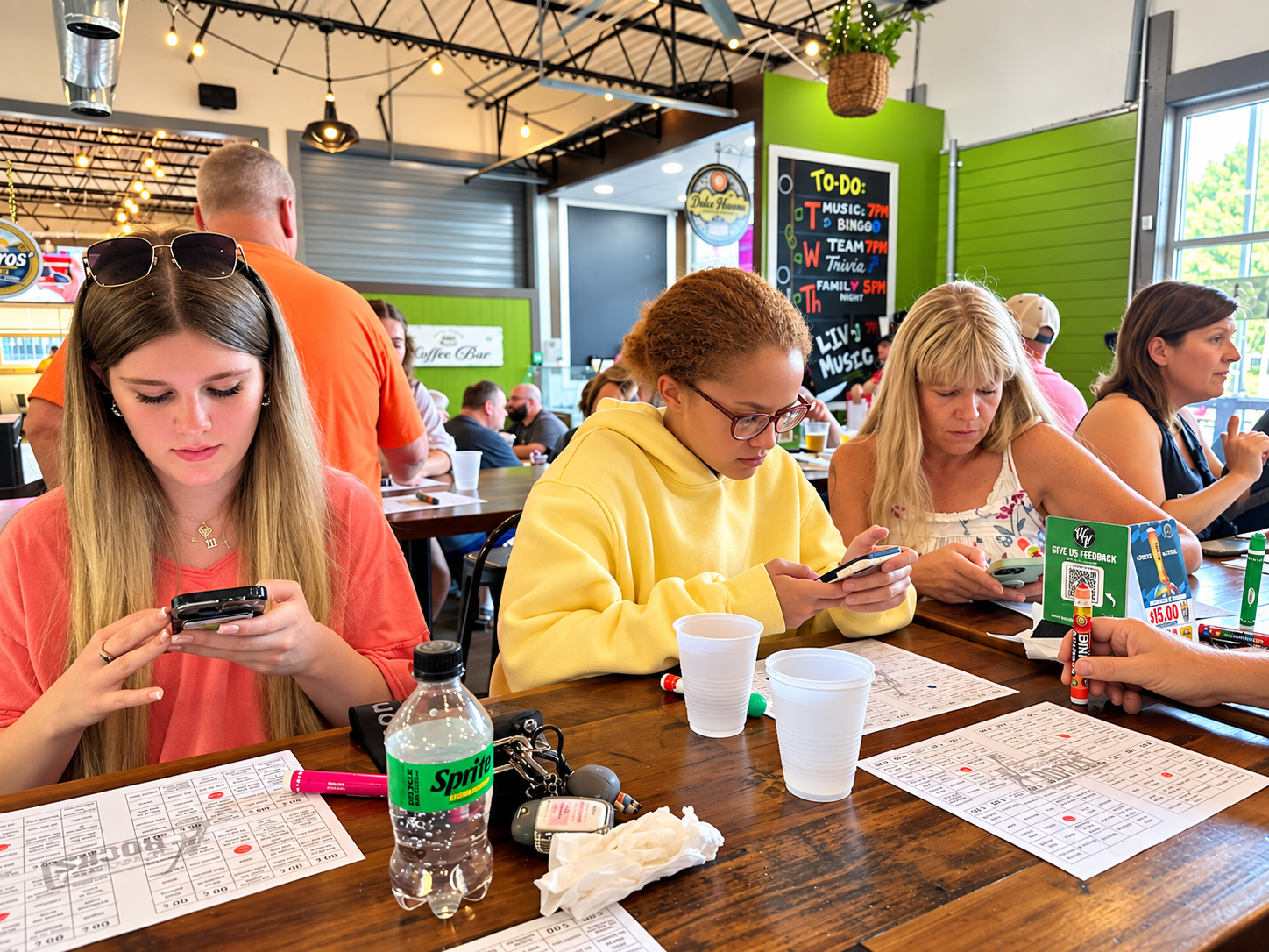 People at a restaurant or cafe, using their phones at a table with menus, drinks, and personal items, with a green wall and blackboard with a to-do list in the background.