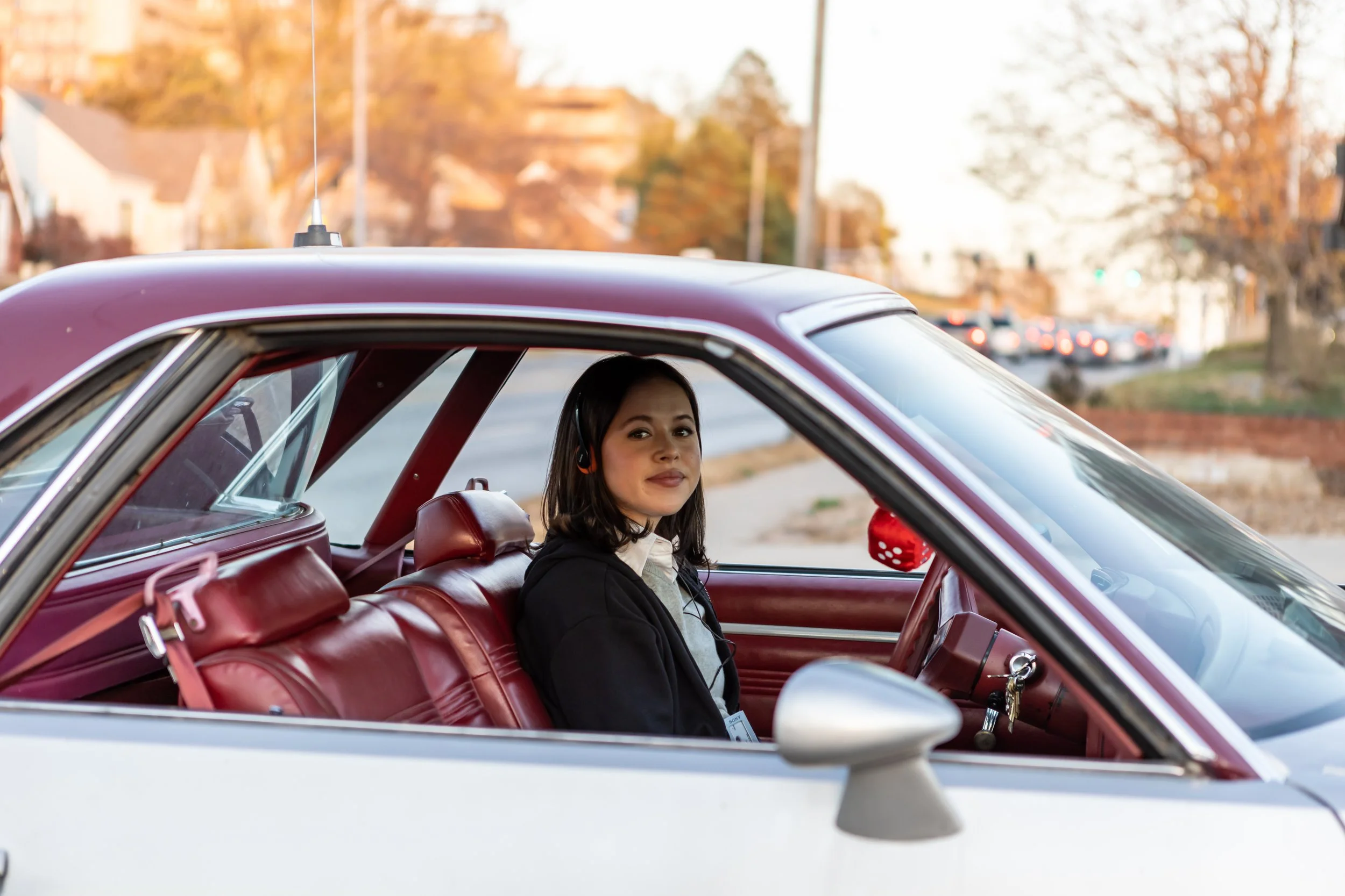 Young woman with dark hair and headphones sitting inside a vintage white car with red interior, parked on a street with autumn trees in the background.