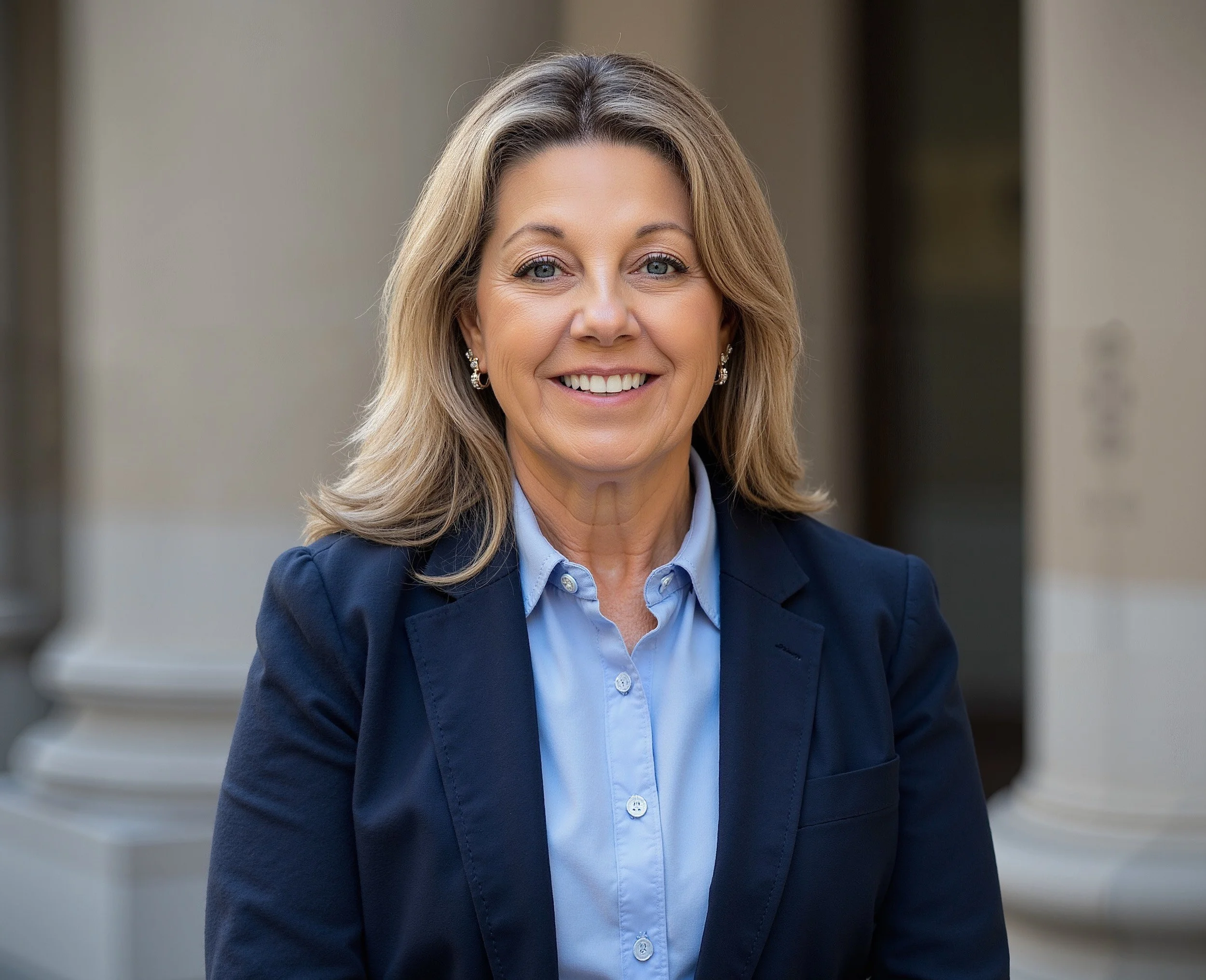 Close-up portrait of a smiling woman with blonde hair, wearing earrings and a navy blazer, standing in front of a building with columns.