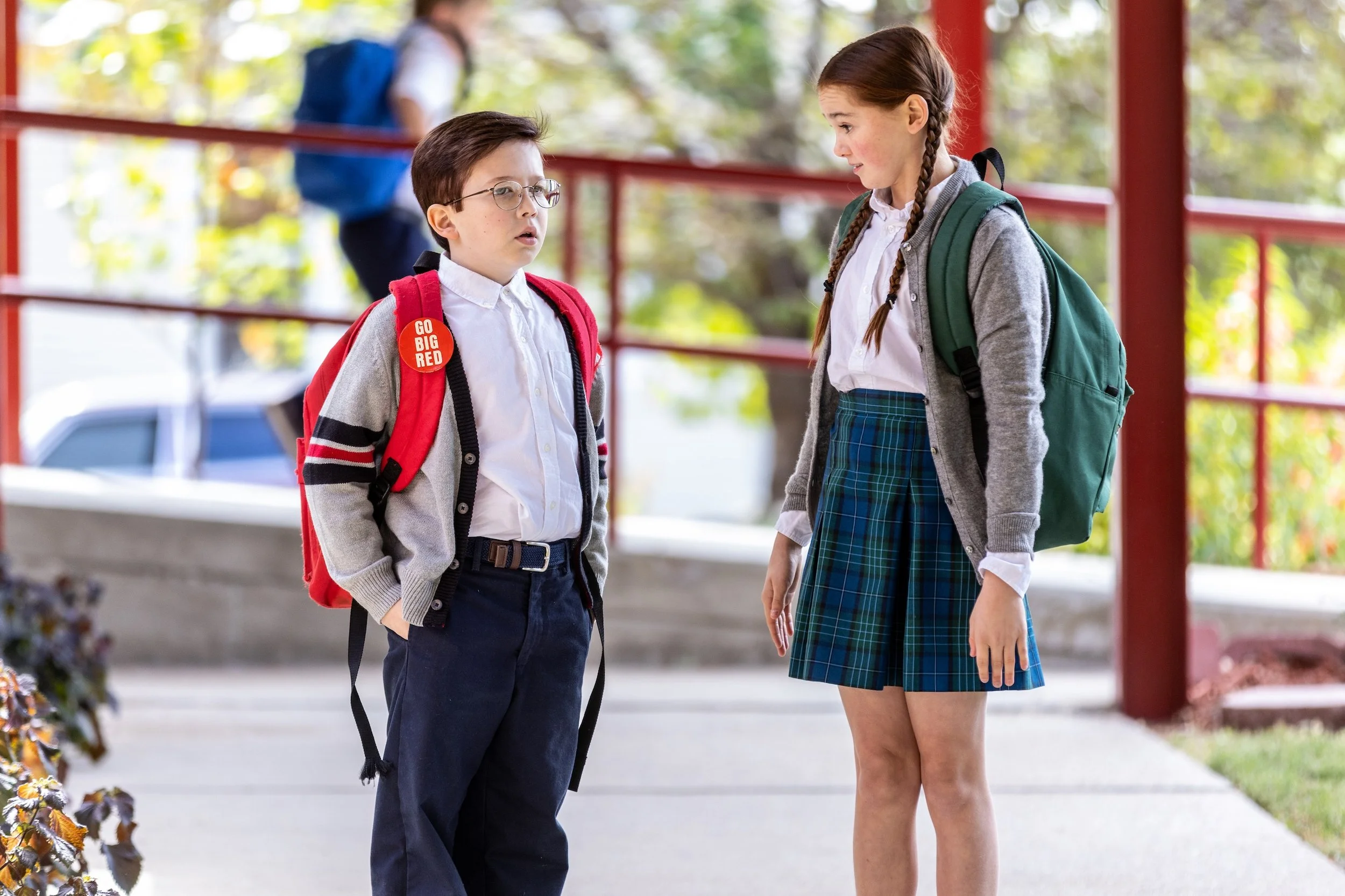 A boy and girl in school uniforms talk outdoors, with backpacks, under a red railing on a sunny day.