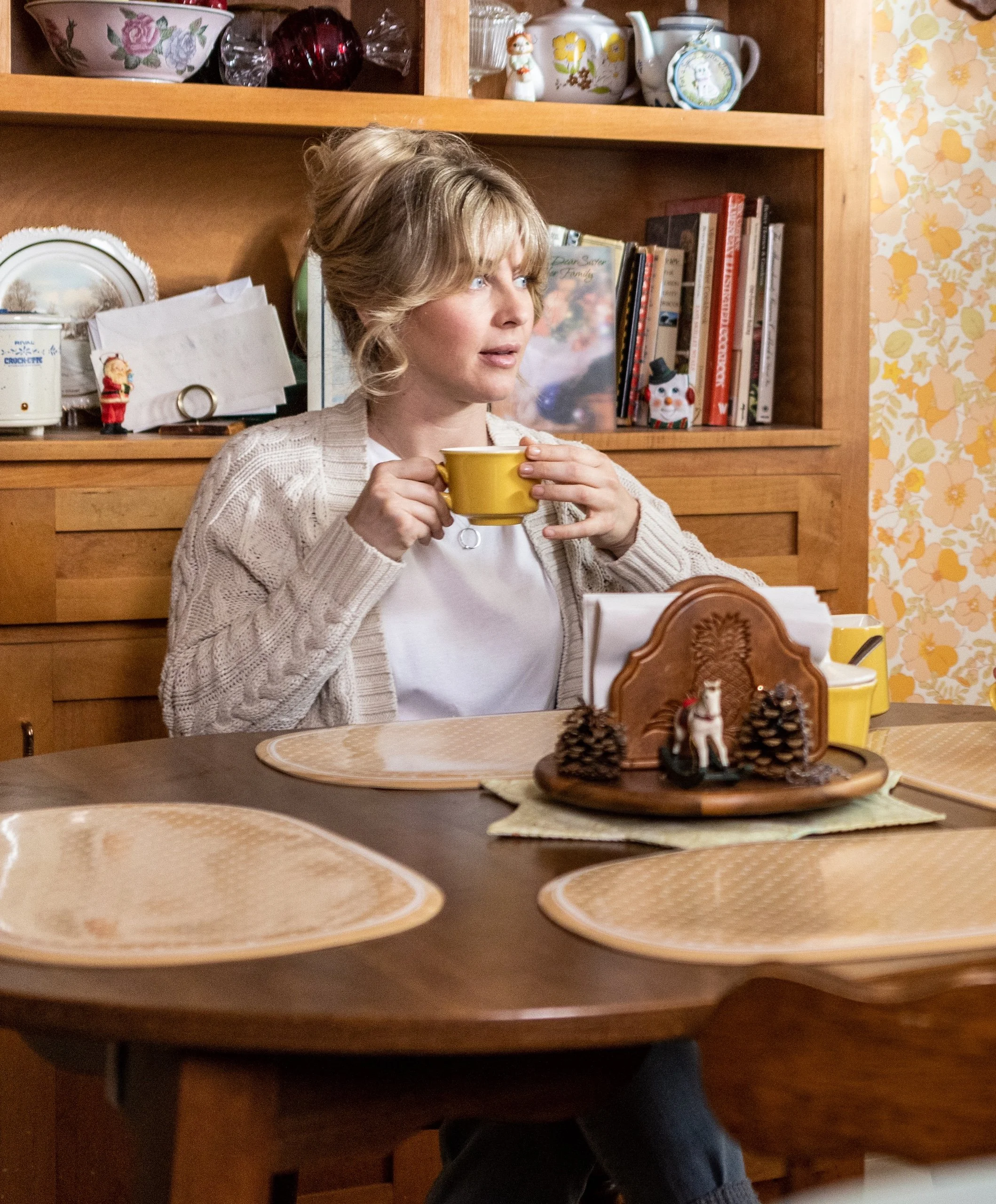 A woman with blonde hair drinking from a yellow cup in a kitchen with wooden cabinets and shelves, decorated with Christmas and holiday items, books, and decorative plates on the wall.