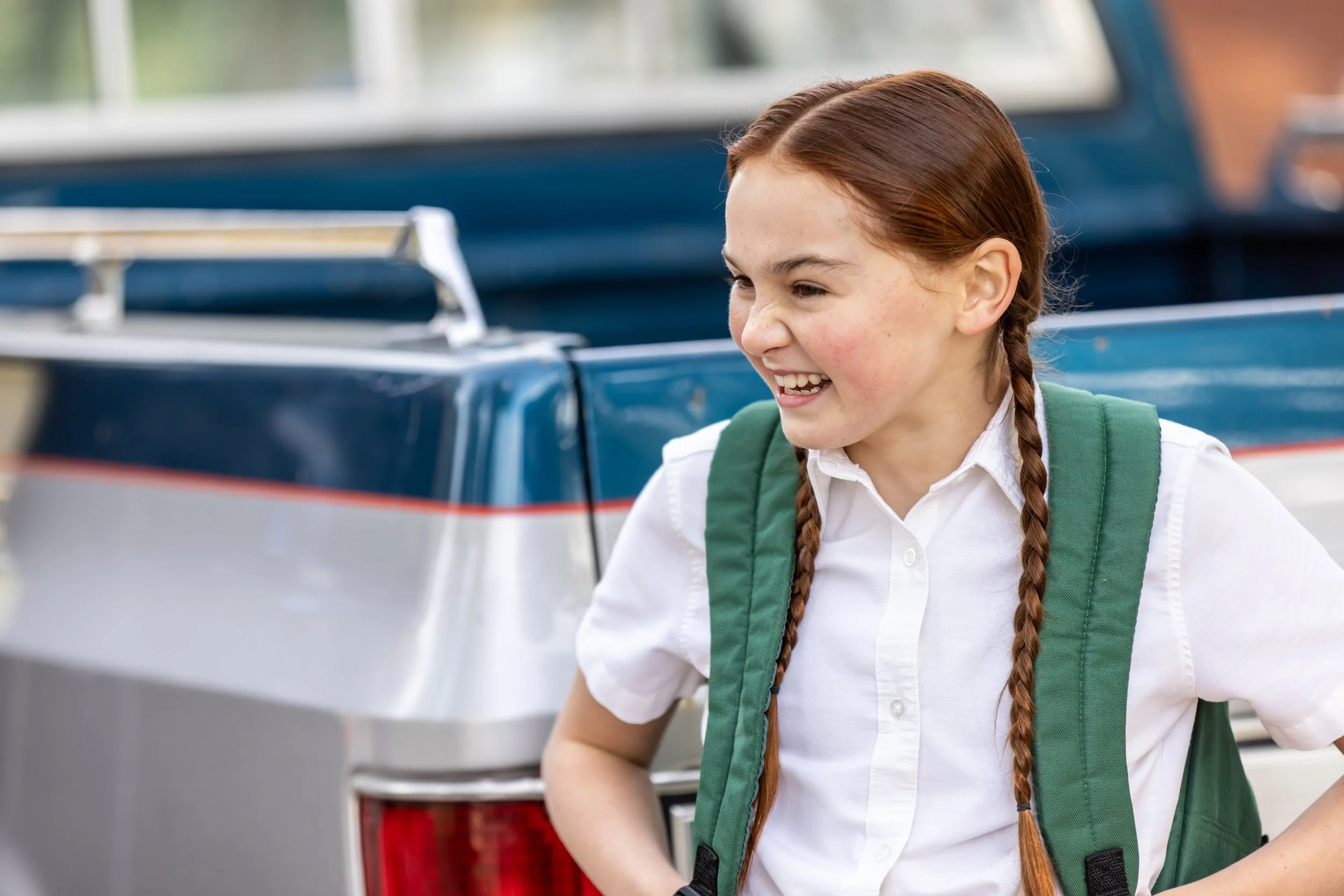 A young girl with red hair in braids, wearing a white shirt and green backpack, smiling and standing next to a blue vintage car.