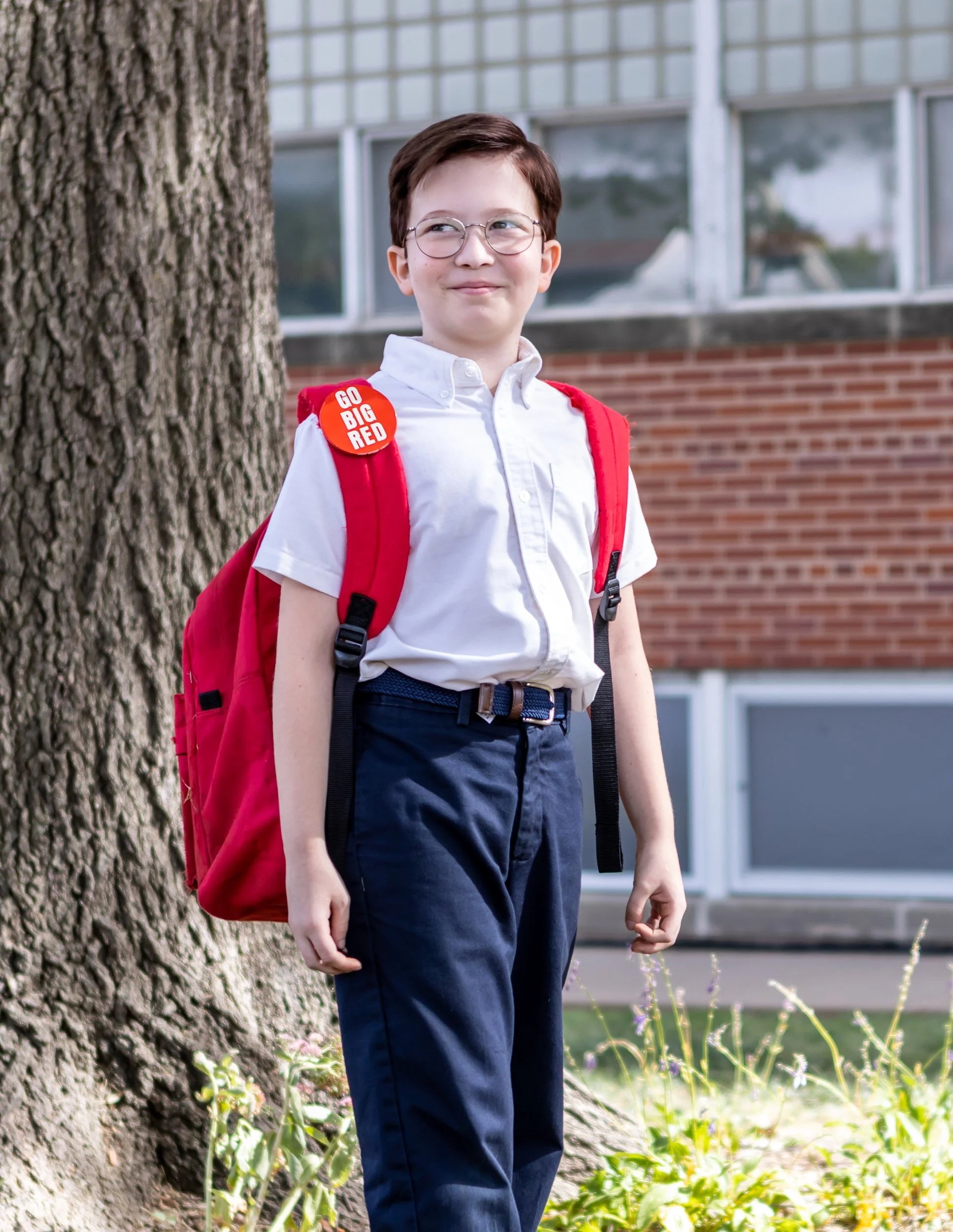 A young boy with short brown hair and glasses, wearing a white shirt and dark pants, standing outside near a tree with a red backpack. It is daytime, and there is a brick building with large windows in the background. The boy has a button on his shirt that says "Go Big Red."