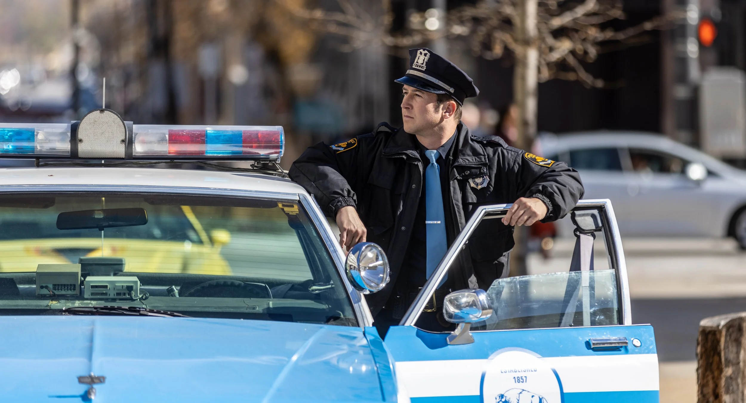 A police officer in uniform leaning on a sheriff's patrol car, looking to the side, on a city street.