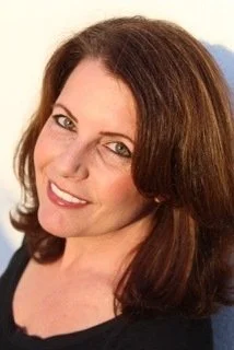 A woman with shoulder-length brown hair smiling at the camera, wearing a black top, with a blurred outdoor background.