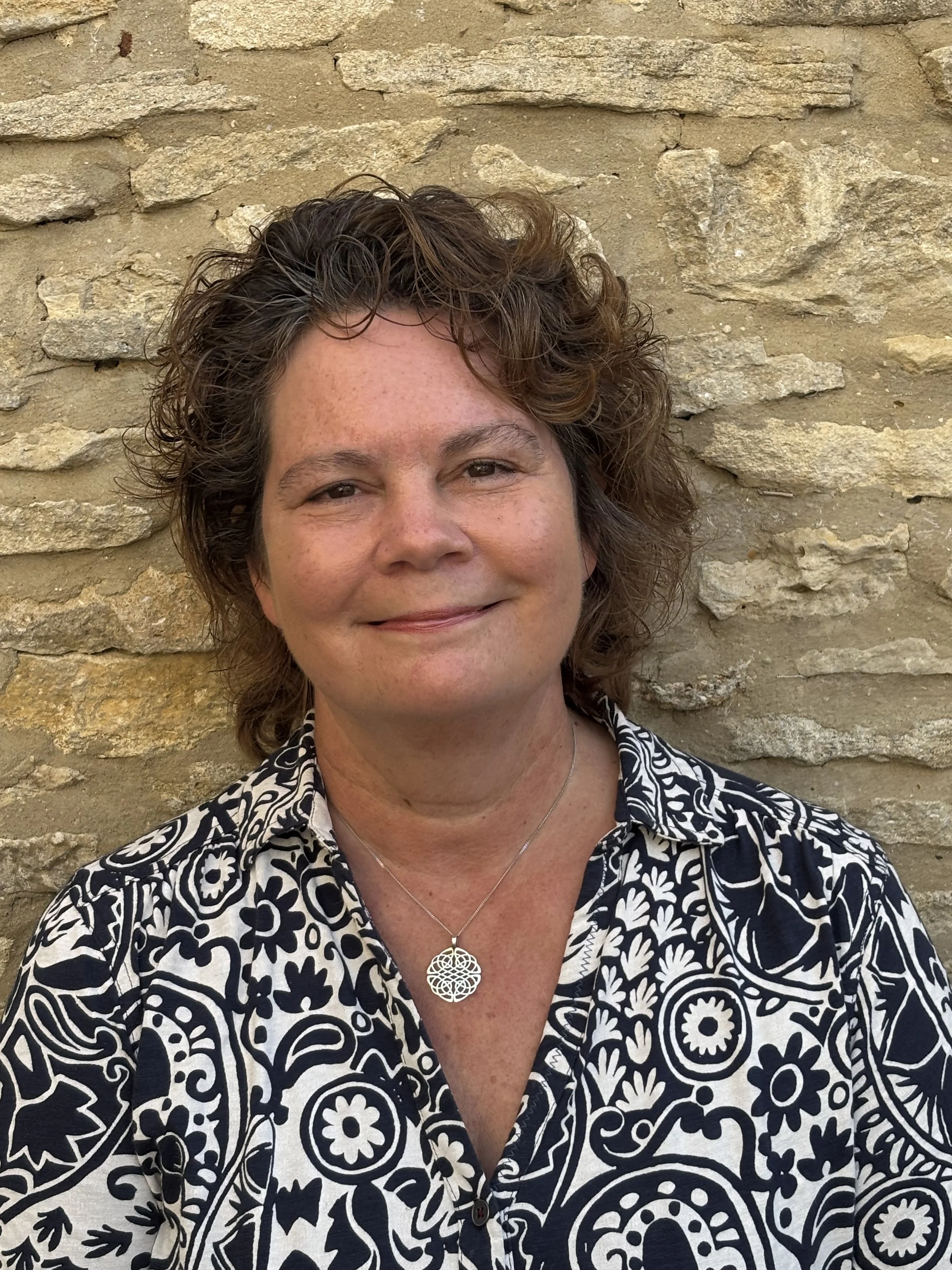 Close-up of a woman in front of a stone wall, wearing a black-and-white patterned shirt and a silver necklace with an ornate pendant, smiling slightly.