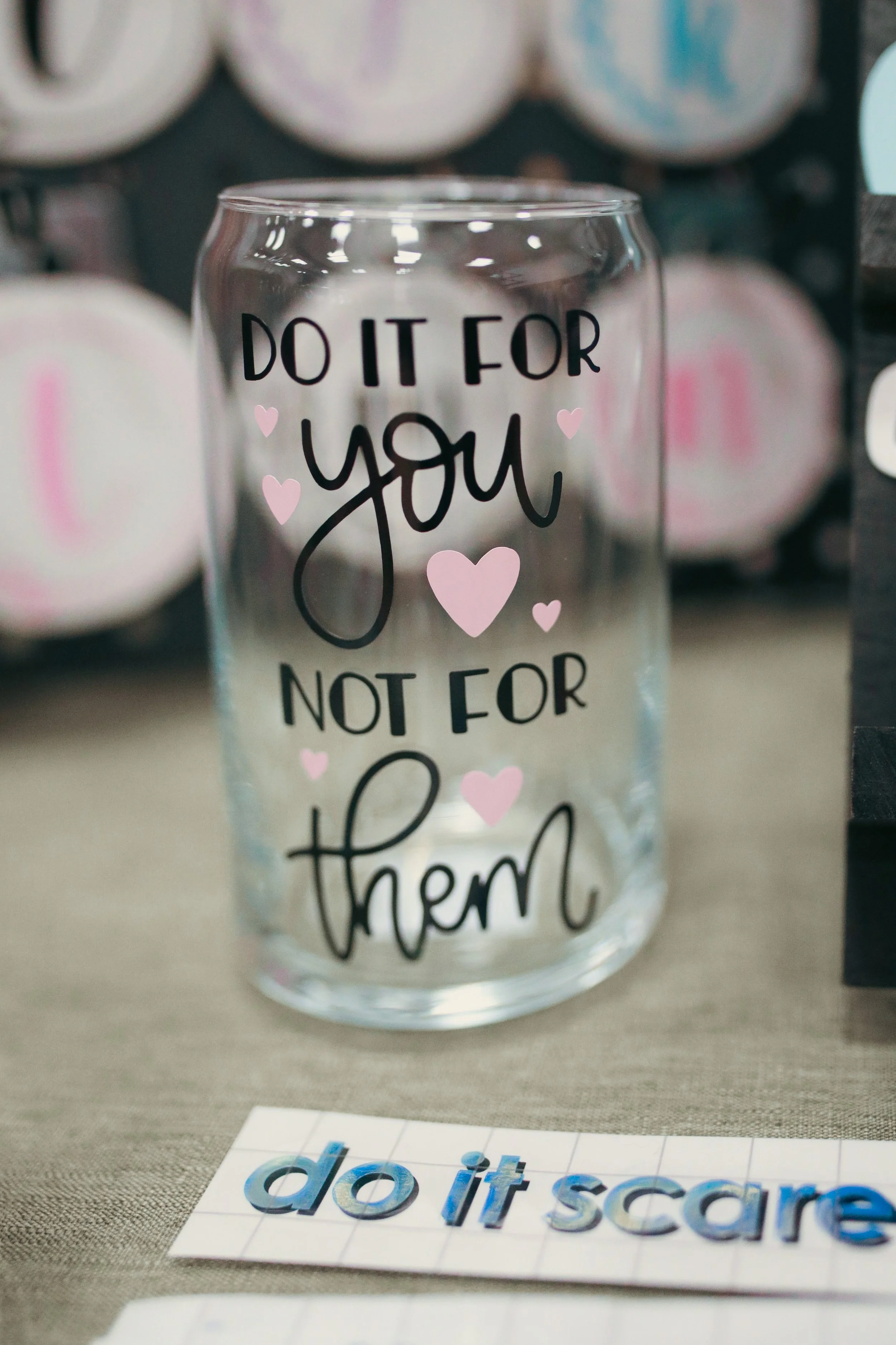 Decorative glass with the phrase 'Do it for you, not for them' surrounded by pink hearts, placed in front of a background of rolls of toilet paper and a sheet of stop stickers reading 'do it scare'.