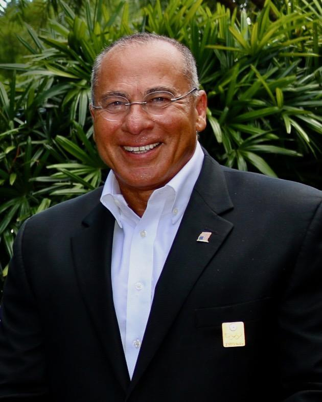 Dr. Nabil El-Hag, smiling, in a dark suit and open-collared blue shirt, outdoors in front of tropical greenery