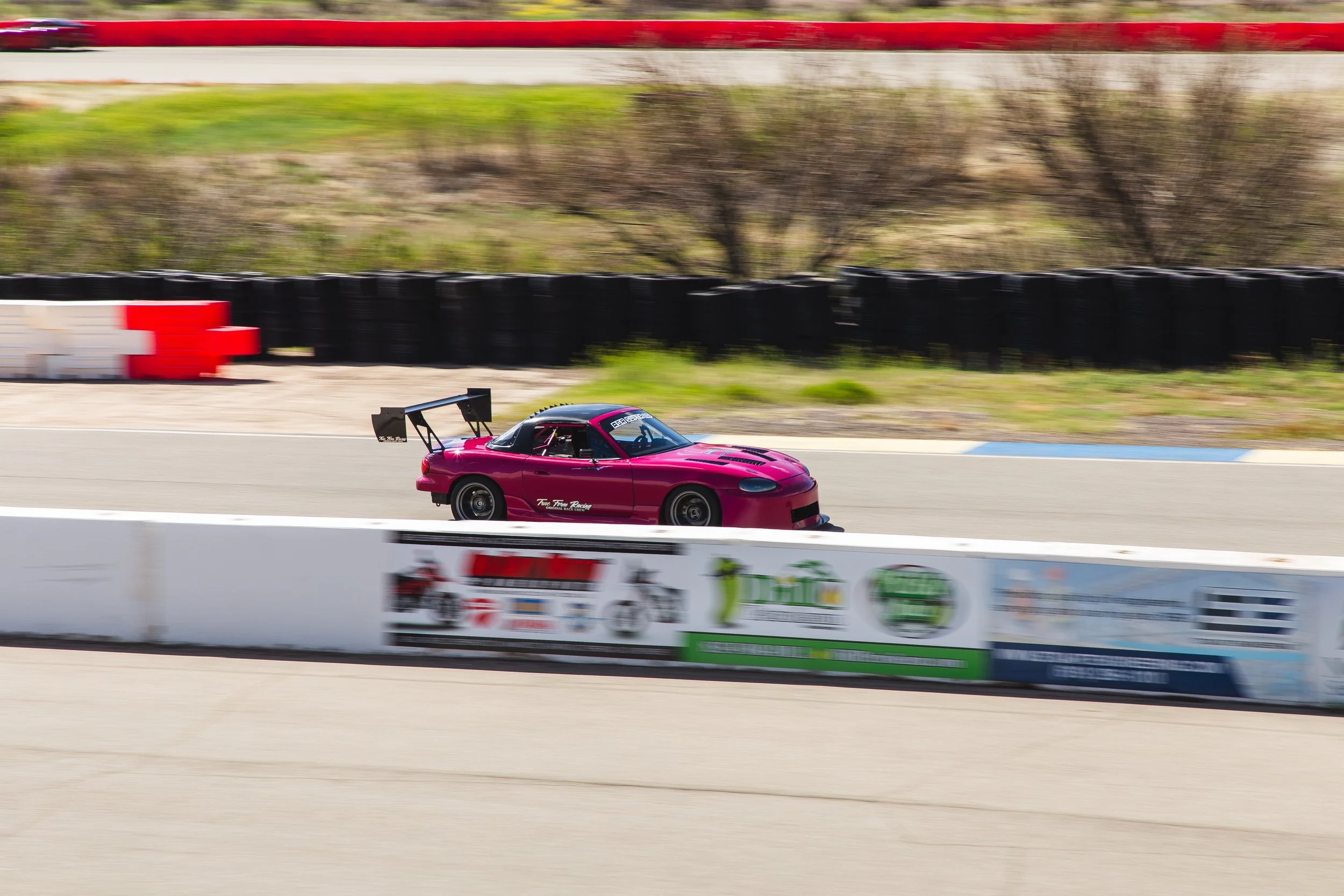 A red race car on a racing track with tire barriers and advertising banners in the background.