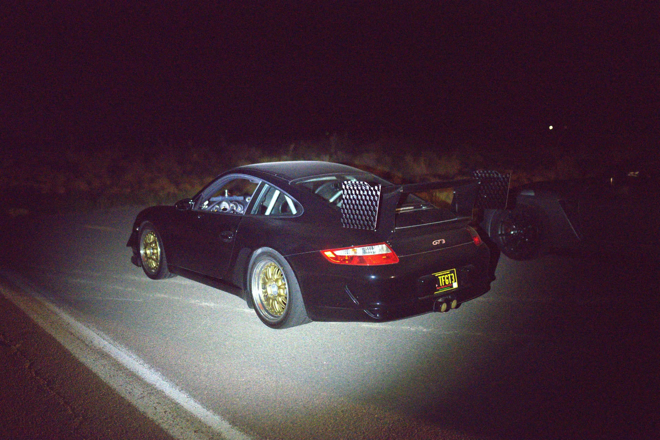 Black Porsche race car with gold wheels parked on the side of the road at night, with the driver's side door open and racing decals on the rear wing.