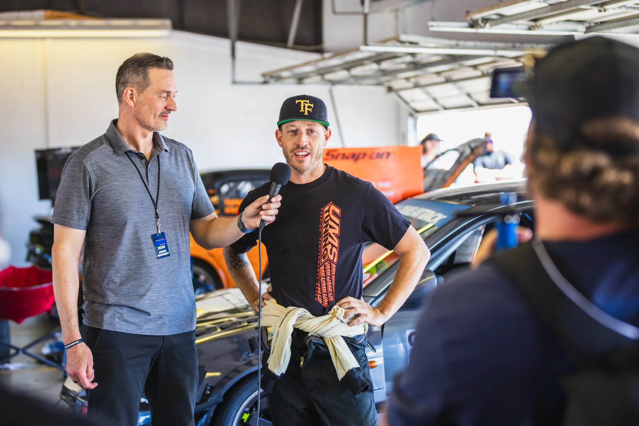 Three men are in a garage with race cars. One man is holding a microphone and two are talking, one wearing a black cap with yellow logo and a racing suit tied around his waist.