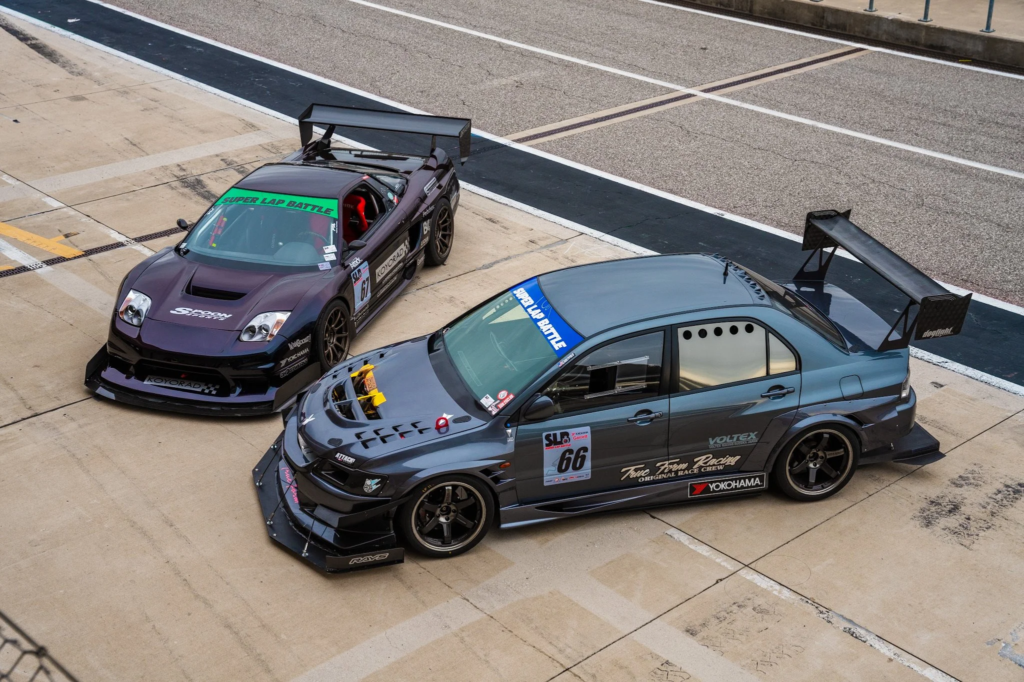 Two race cars parked side by side on a racetrack pit lane, one black with aerodynamic spoilers and decals, the other dark gray with a large rear wing and racing stickers.