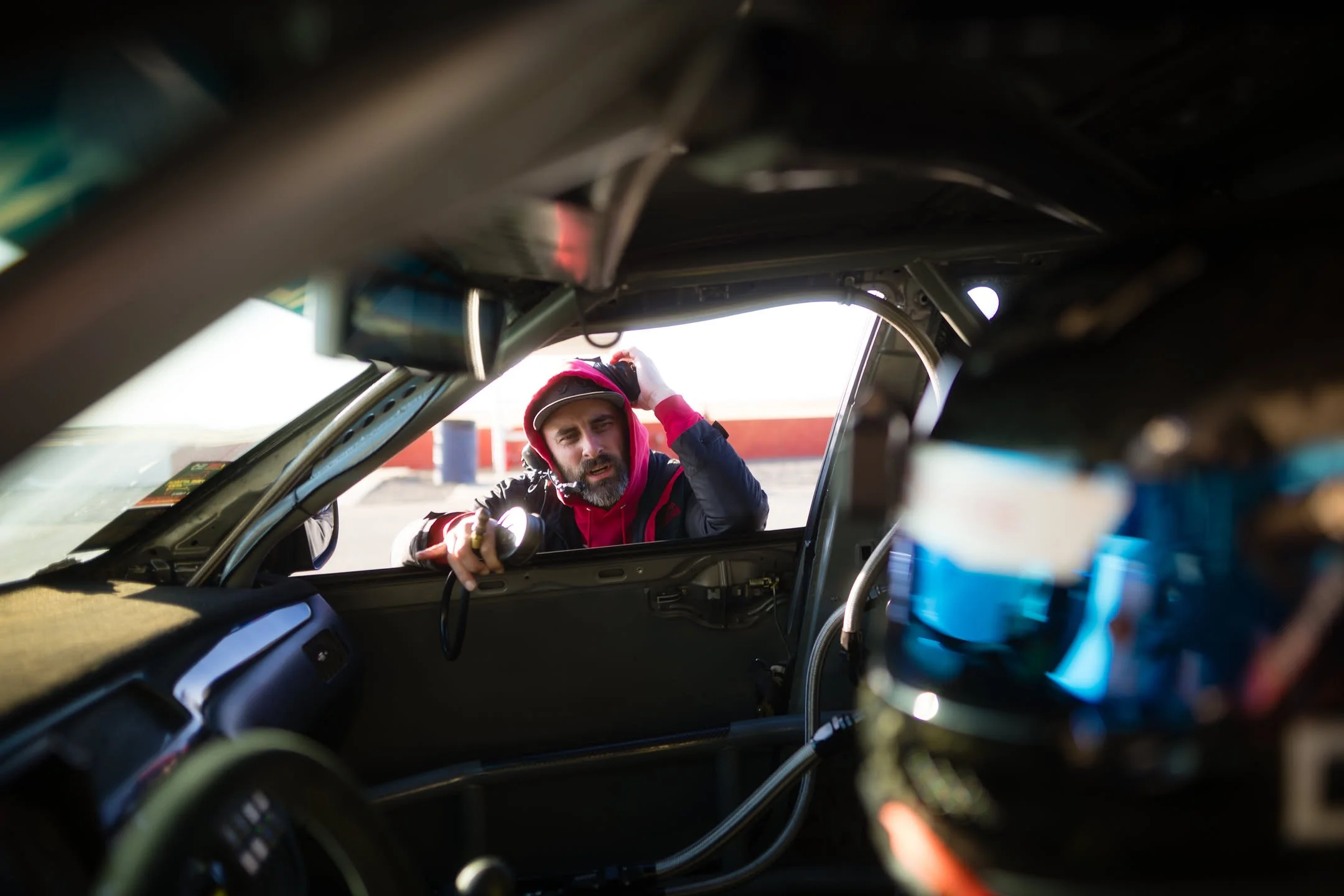 A man with a beard and red hoodie is working on a vehicle, seen from inside the car looking out through the open door, with a focus on the man and the interior of the car.