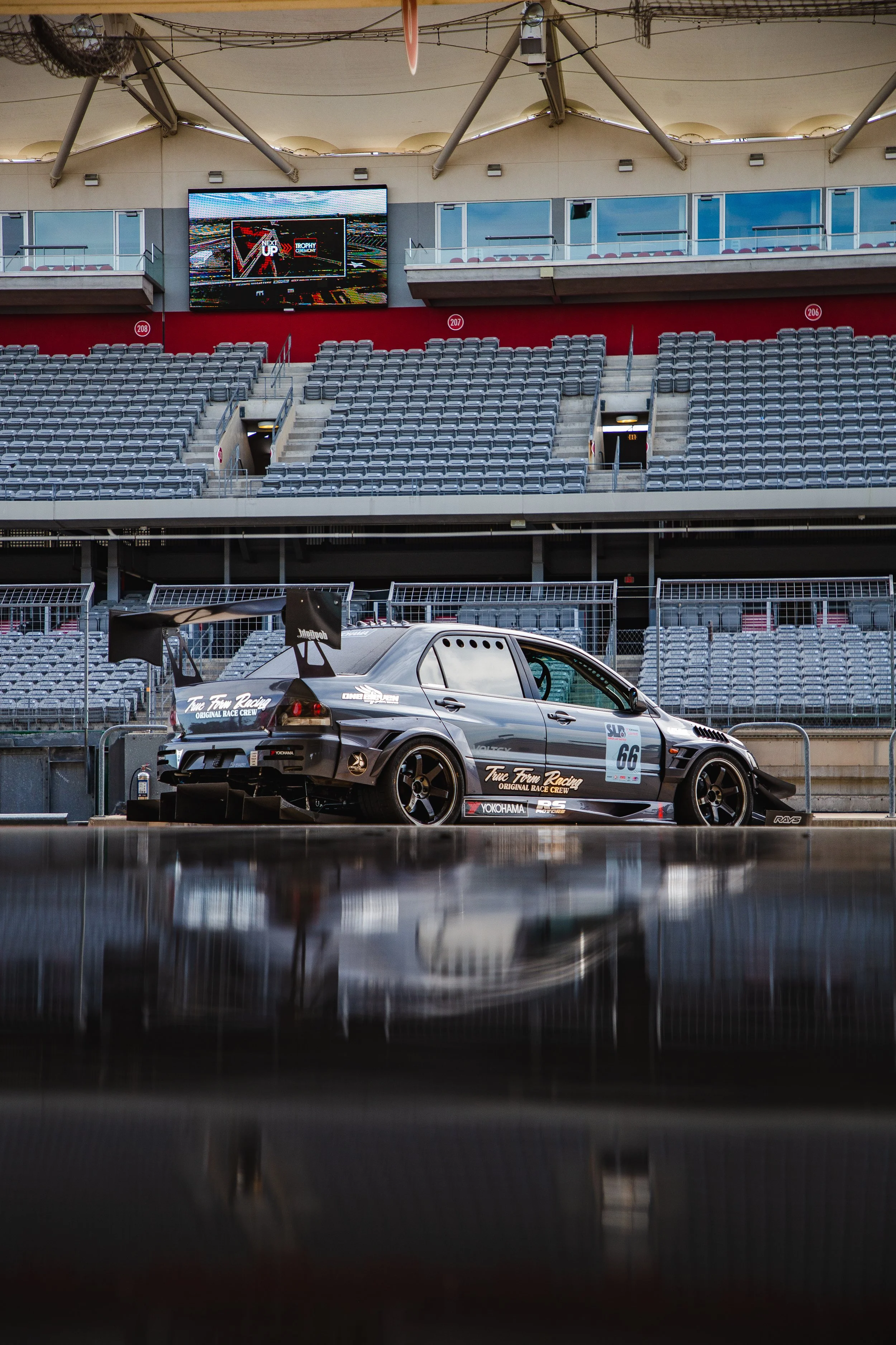 A race car with sponsorship logos parked on a race track at a stadium. The background shows empty seats and a large digital screen displaying advertisements.