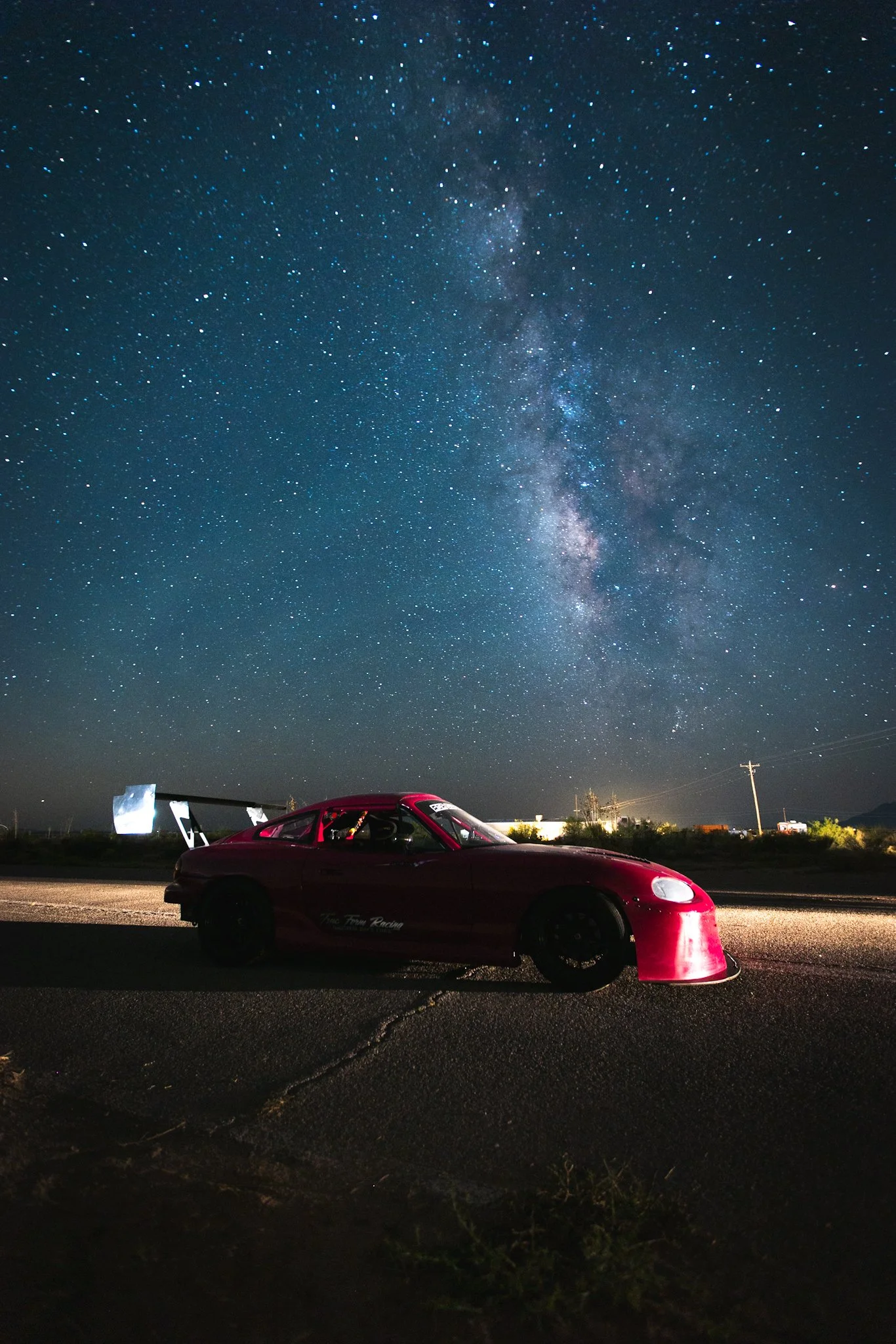 A red sports car parked on the side of a road with a star-filled night sky and the Milky Way galaxy overhead.