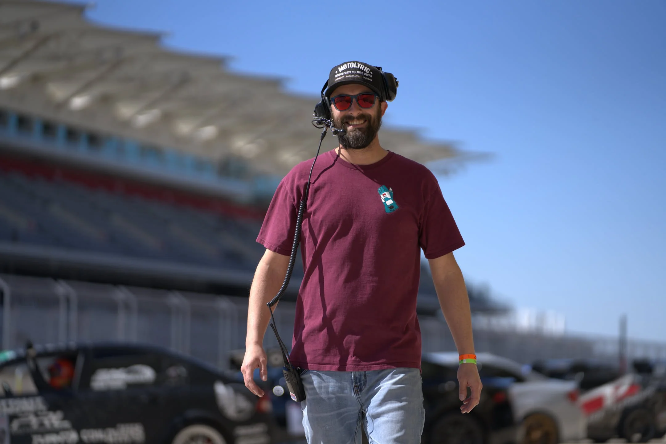 A man with sunglasses, a beard, and a maroon t-shirt smiling while walking outdoors on a sunny day, with a background of cars and a building.