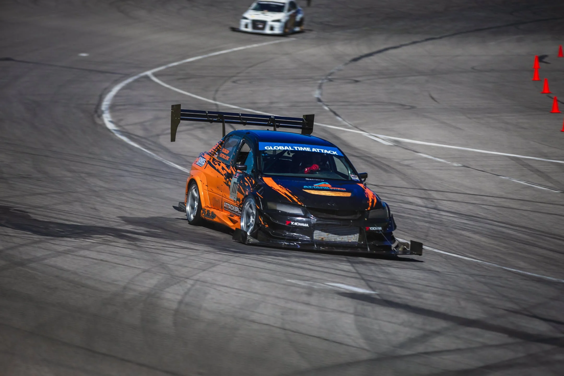 A race car with orange and black livery on a race track, surrounded by orange traffic cones, and a white race car in the background.