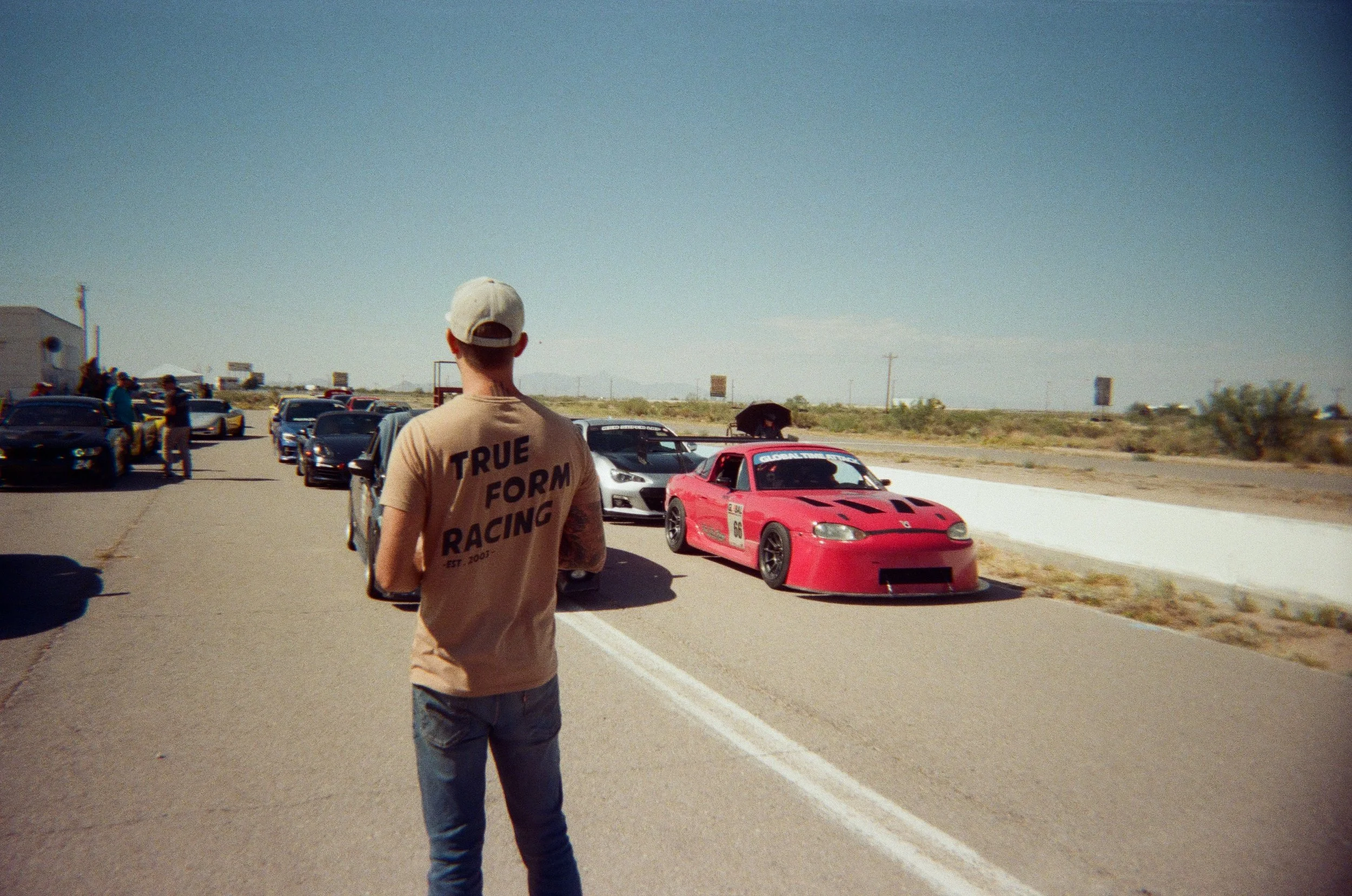 A person wearing a beige hat and a beige T-shirt with black text, standing on a desert highway, with several race cars parked nearby, including a pink race car with the number 66, and other people in the background.