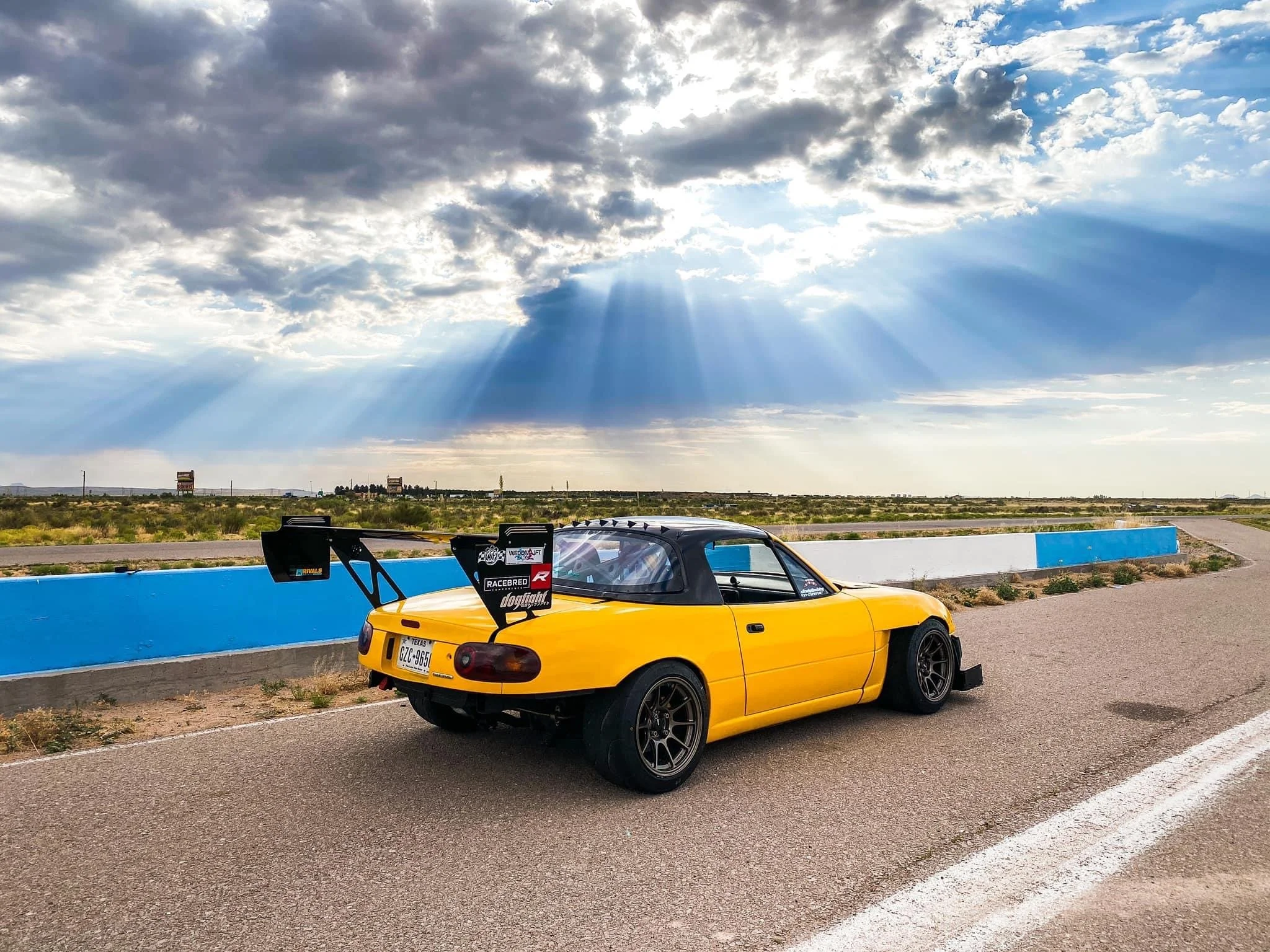 A yellow sports car with racing decals parked on a race track, with a blue barrier and open landscape in the background under a cloudy sky with sun rays breaking through.