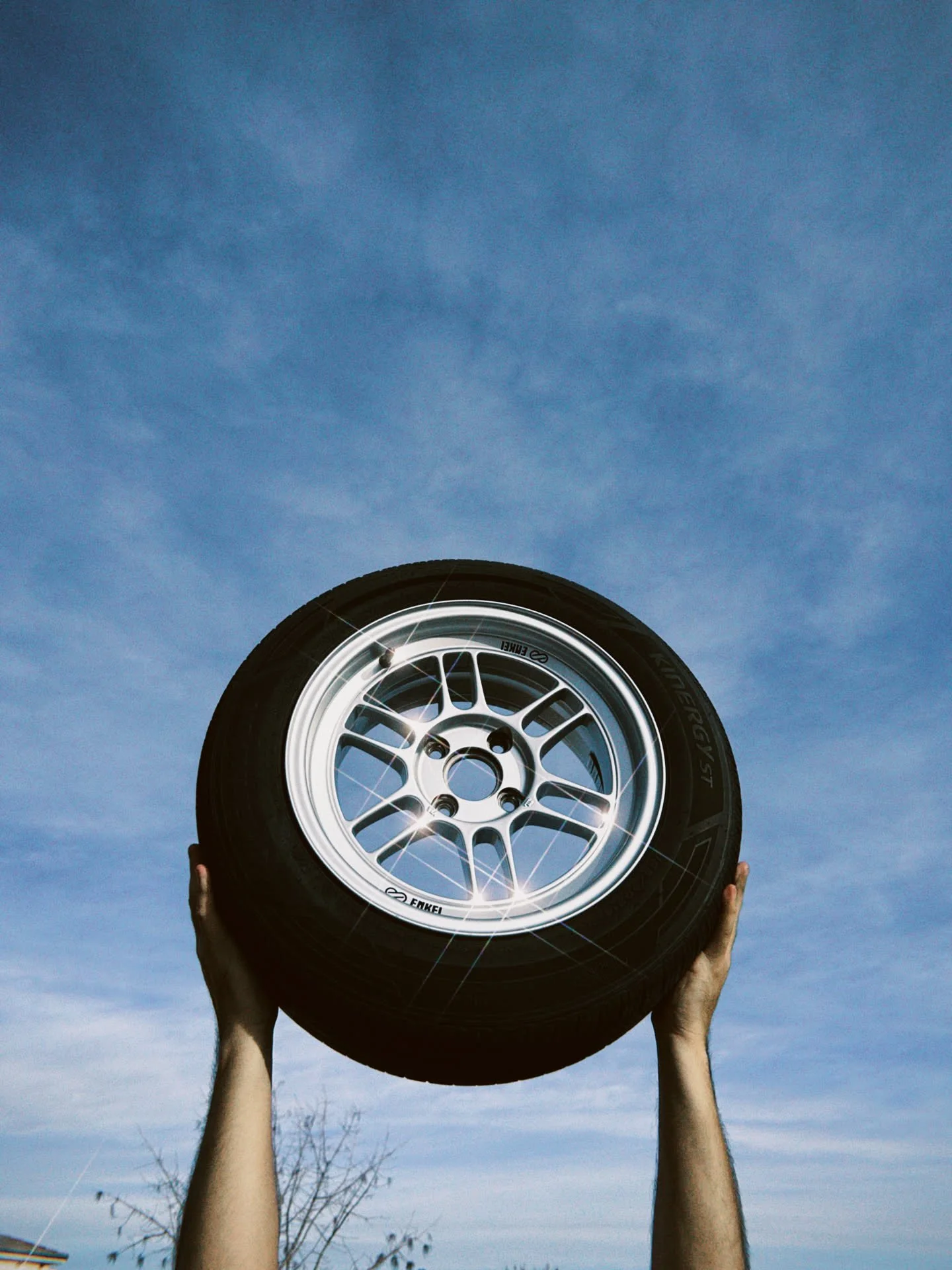 A person holding a car tire with a silver rim up against a bright blue sky with some clouds.