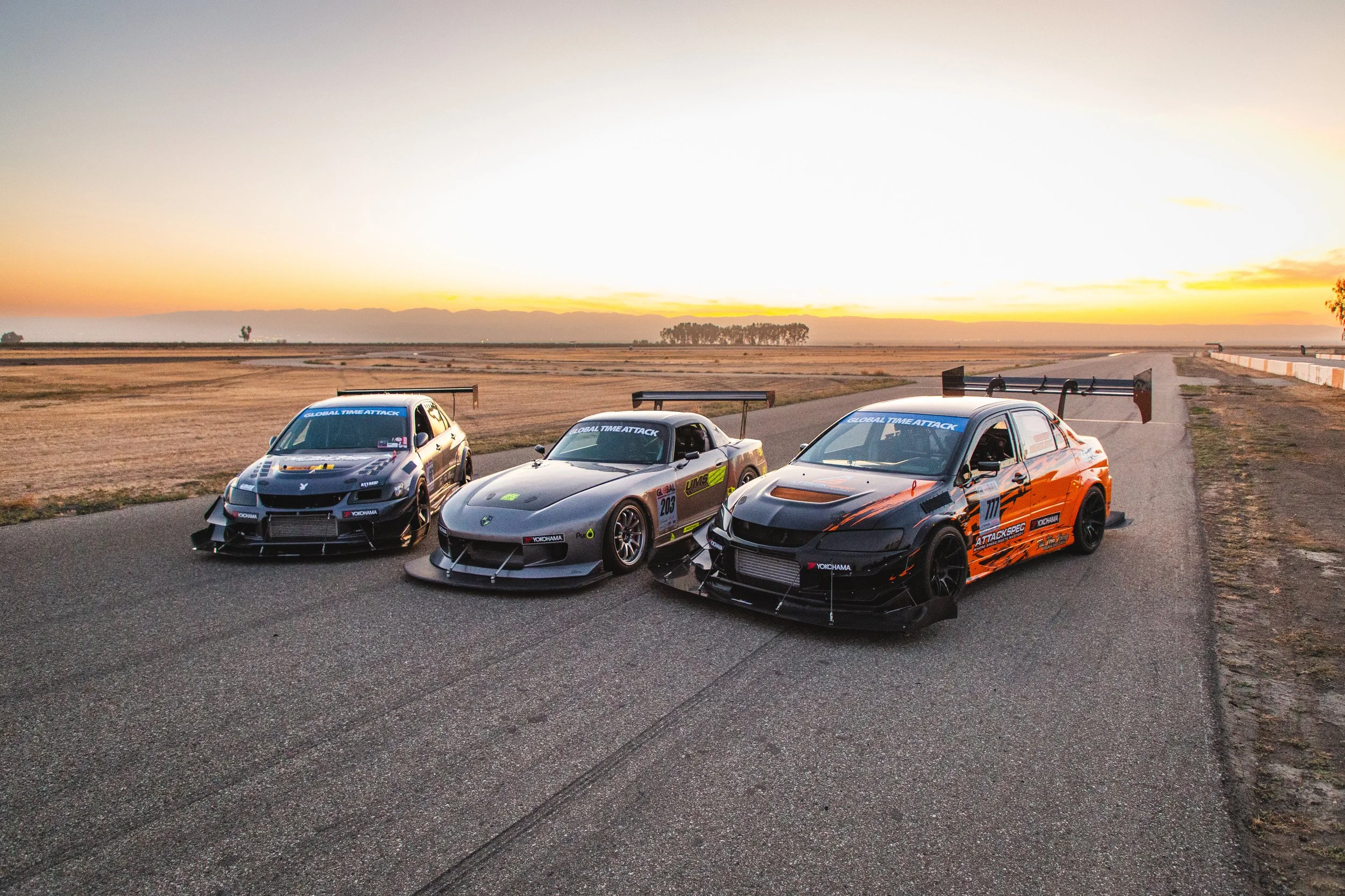 Three race cars parked on a track at sunset, with open fields and mountains in the background.