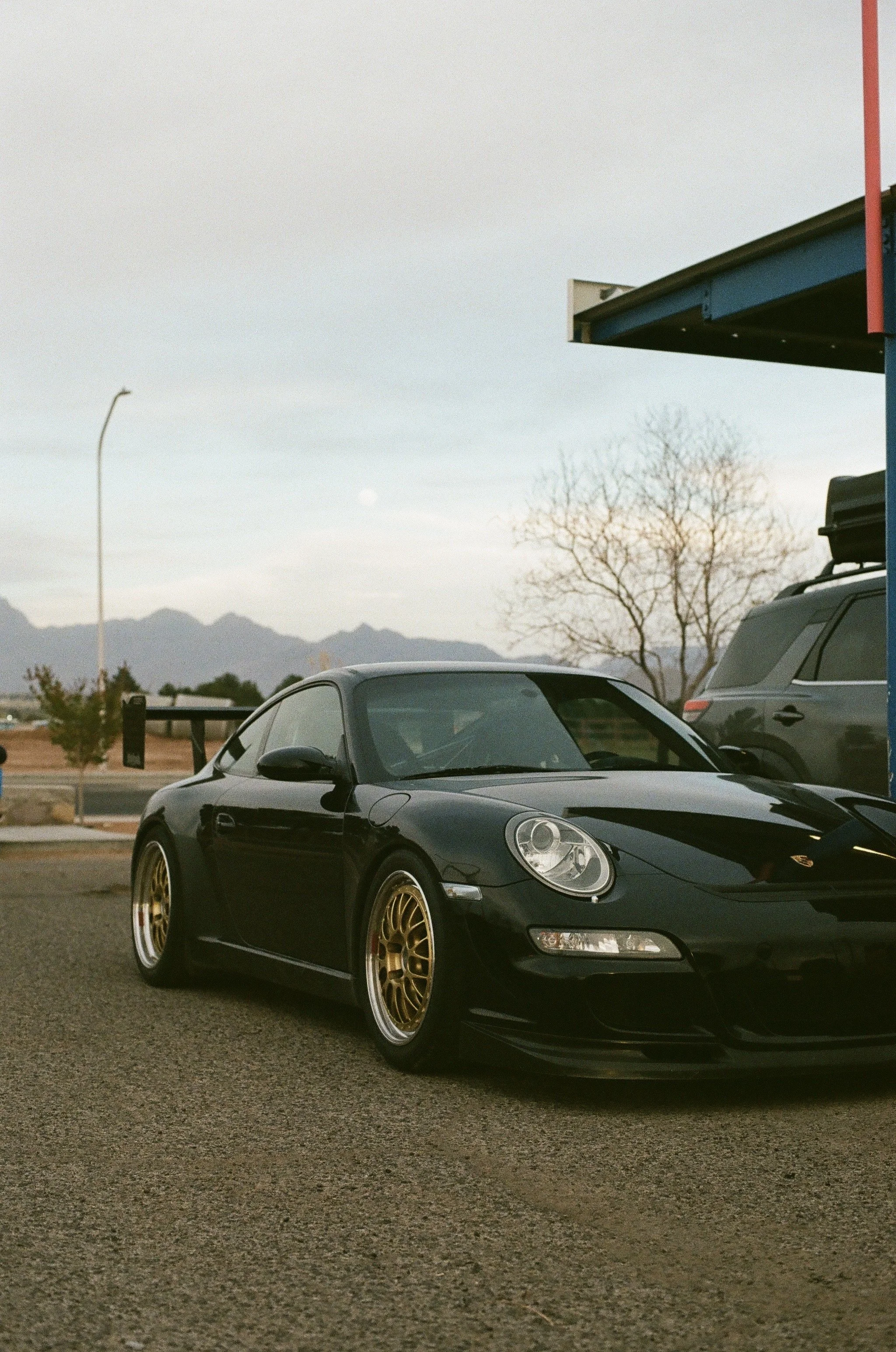 A black sports car with custom gold wheels parked in a parking lot, with mountains in the background and a clear sky.