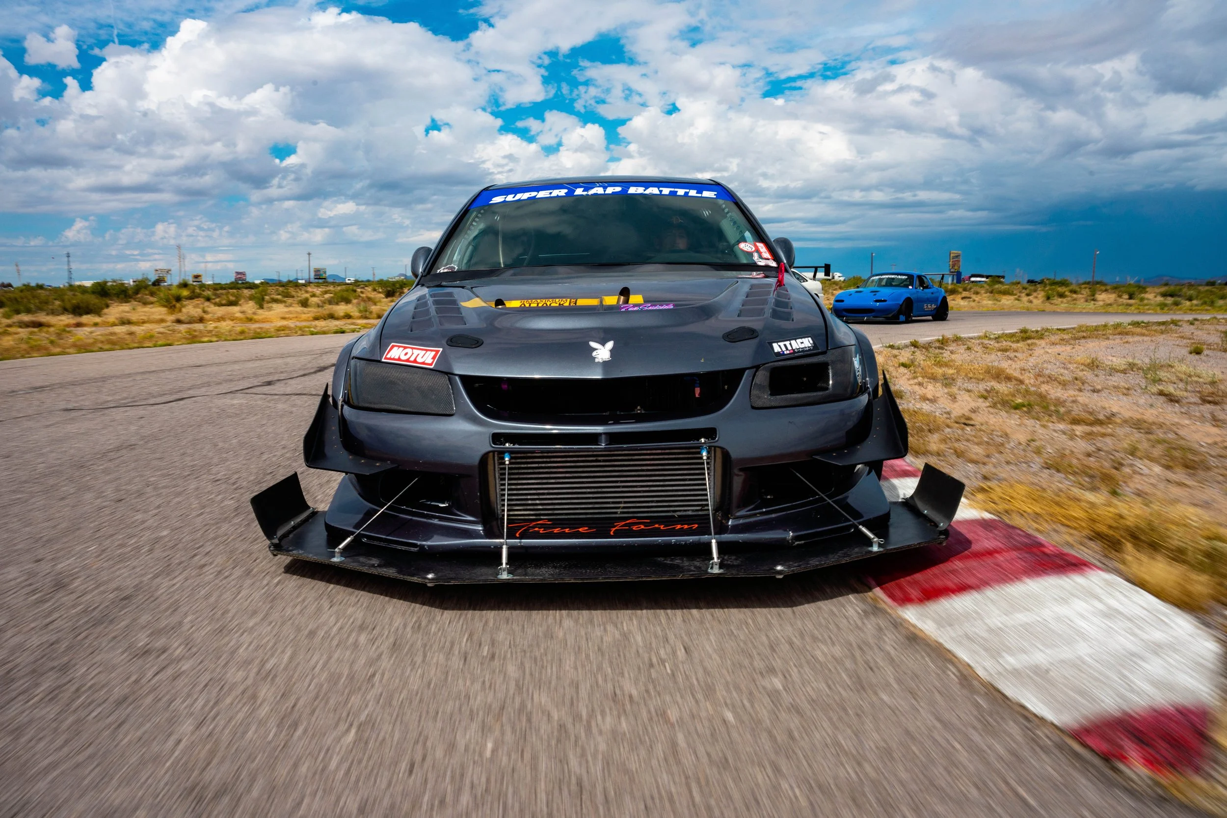Race cars driving on a desert race track under a cloudy sky, with a black car in the foreground featuring decals and a blue car in the background.