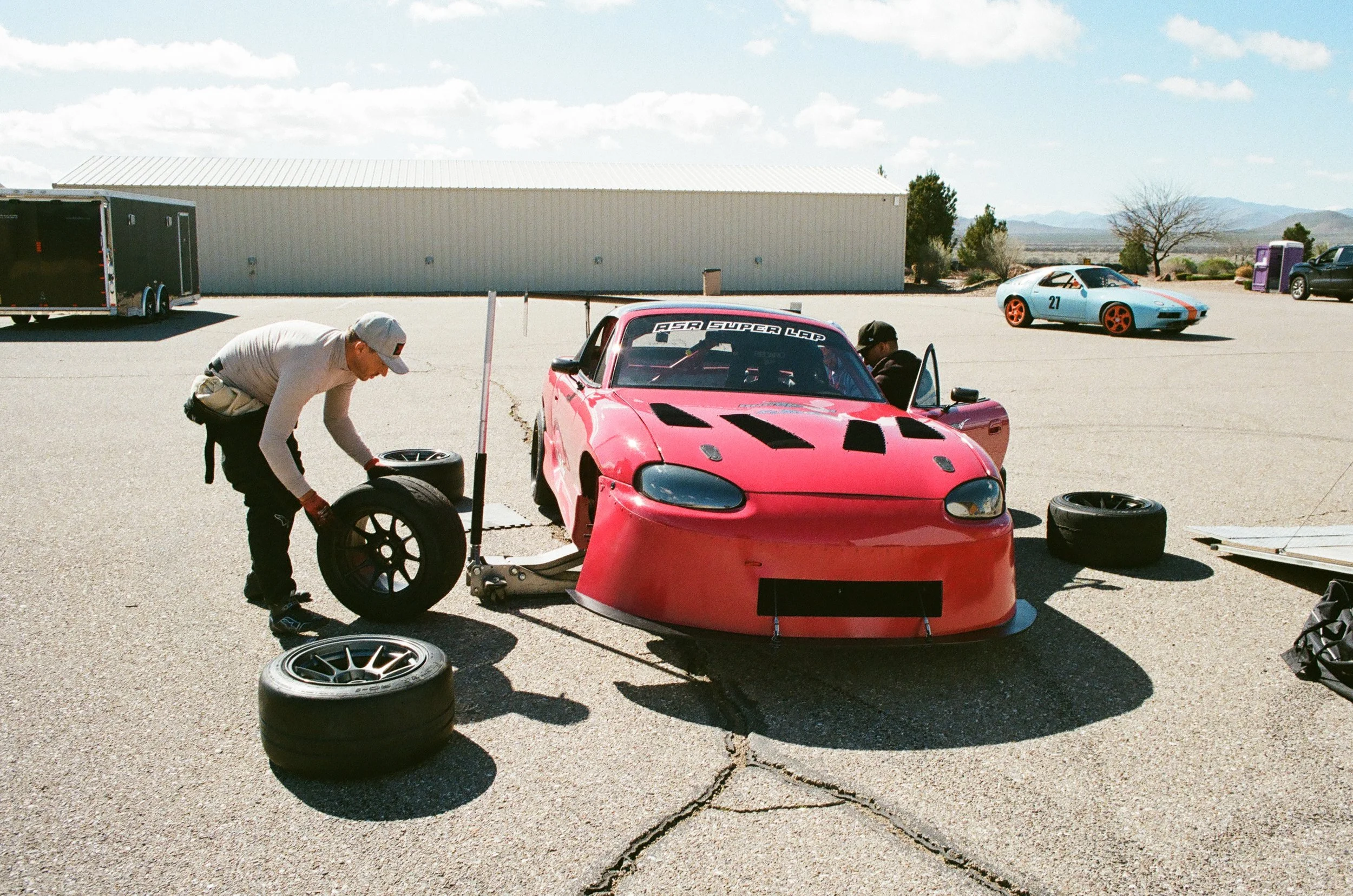 A person working on a pink race car with a crew member nearby, in an open outdoor area. Two other race cars are visible in the background, along with tires, a trailer, and a large metal building. The sky is mostly clear with some clouds.