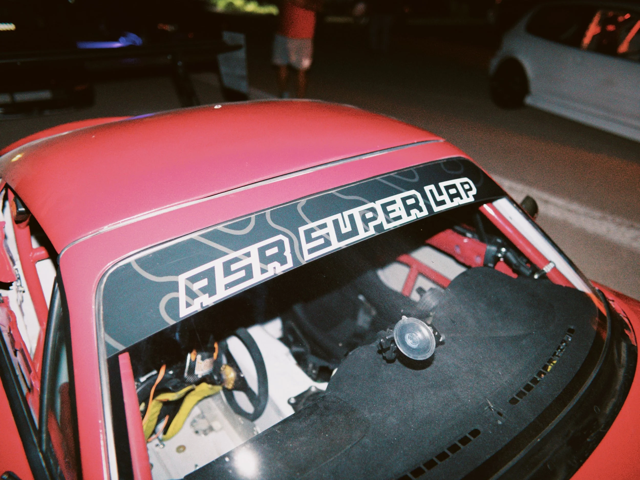 A red remote-controlled car with a black windshield sticker that says 'SUPERLAP', showing the interior components including wiring and engine parts, with a green plant in the background.