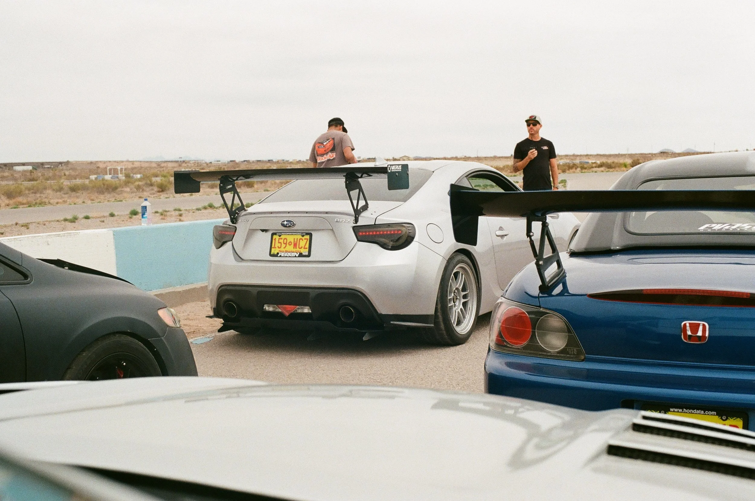 Two men stand next to modified cars with large rear wing spoilers, in a desert-like racing track environment, with multiple vehicles in the background.