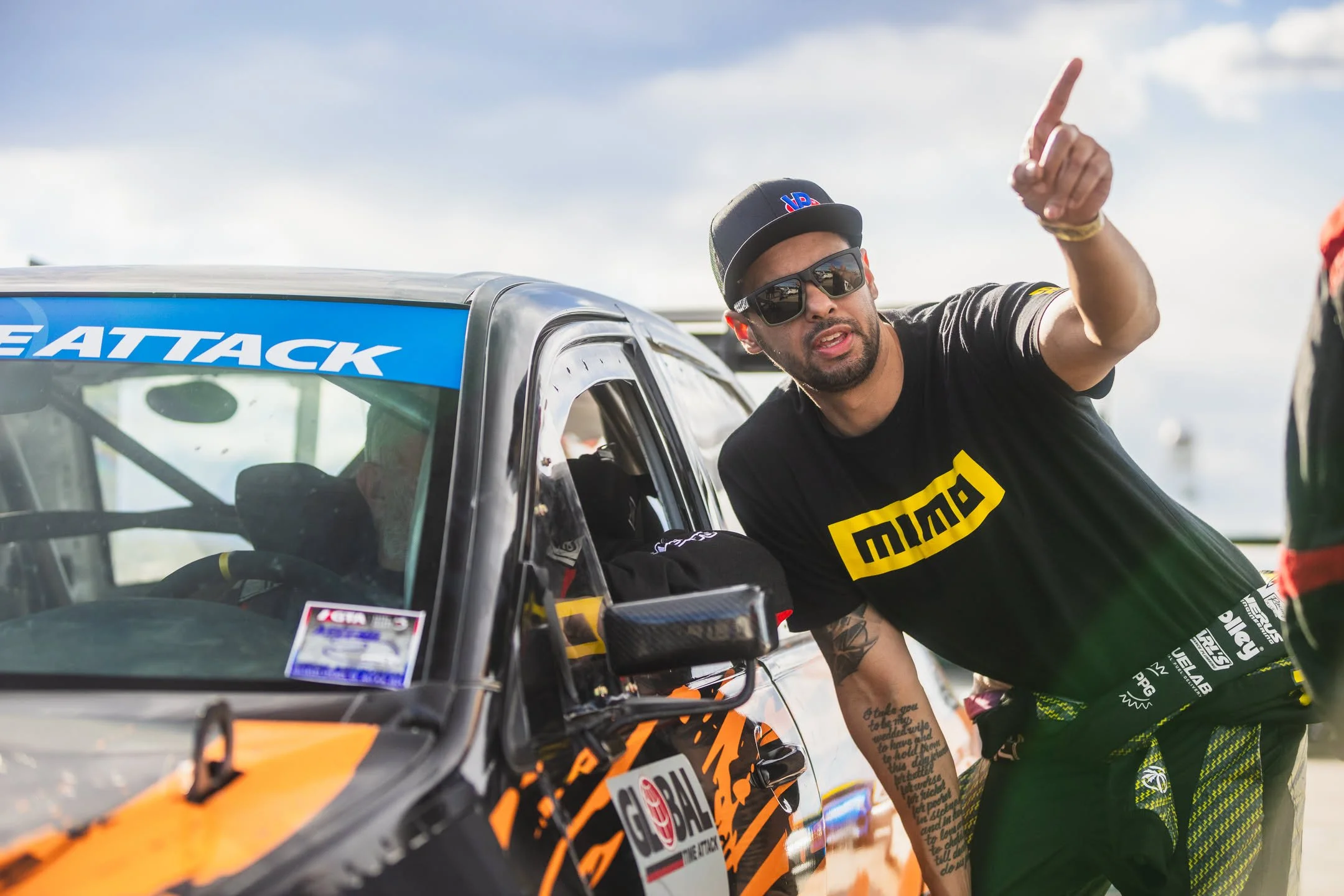 A man wearing sunglasses and a black cap with an American flag and 'HOn' written on it leans out of a black and orange race car, pointing upwards, with a woman in green clothing next to him at what appears to be a racing event.