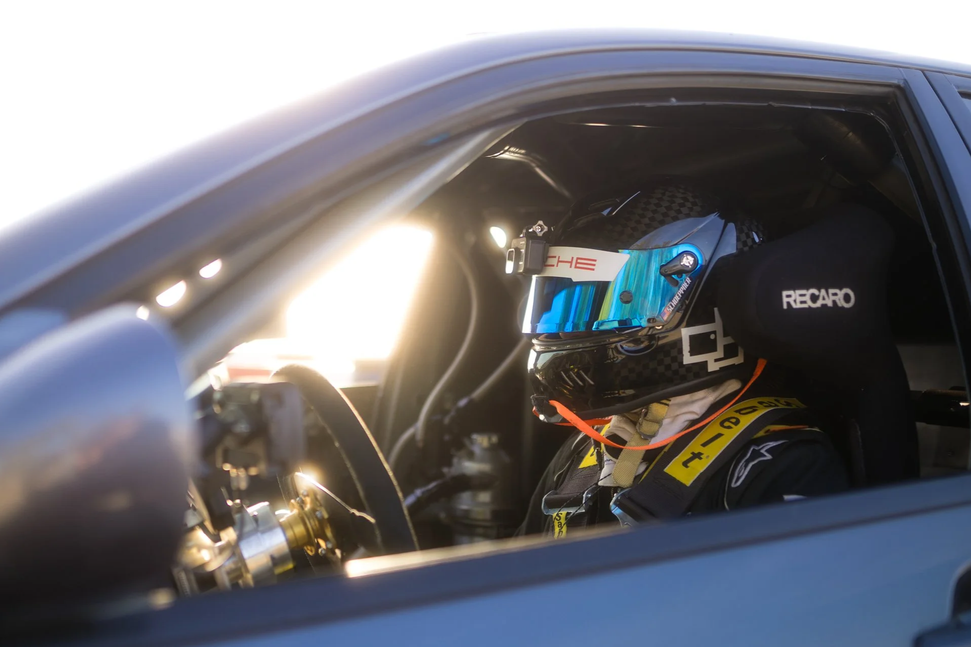 Close-up of a race car driver wearing a helmet and racing suit, seated inside a race car, with sunlight shining through the window.